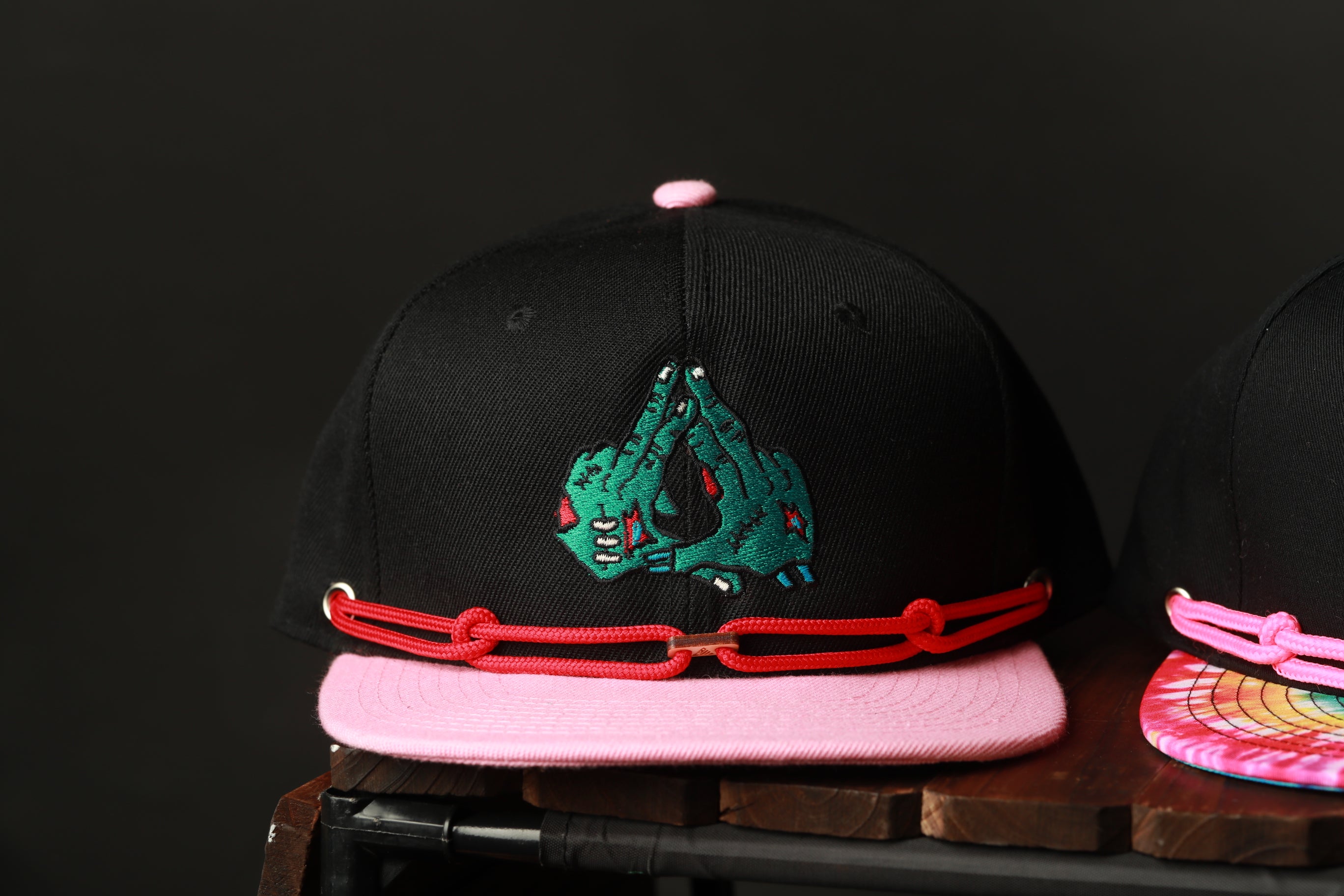 A black baseball cap with a pink brim and a graphic of a green hand on the front, sitting on a wooden surface.