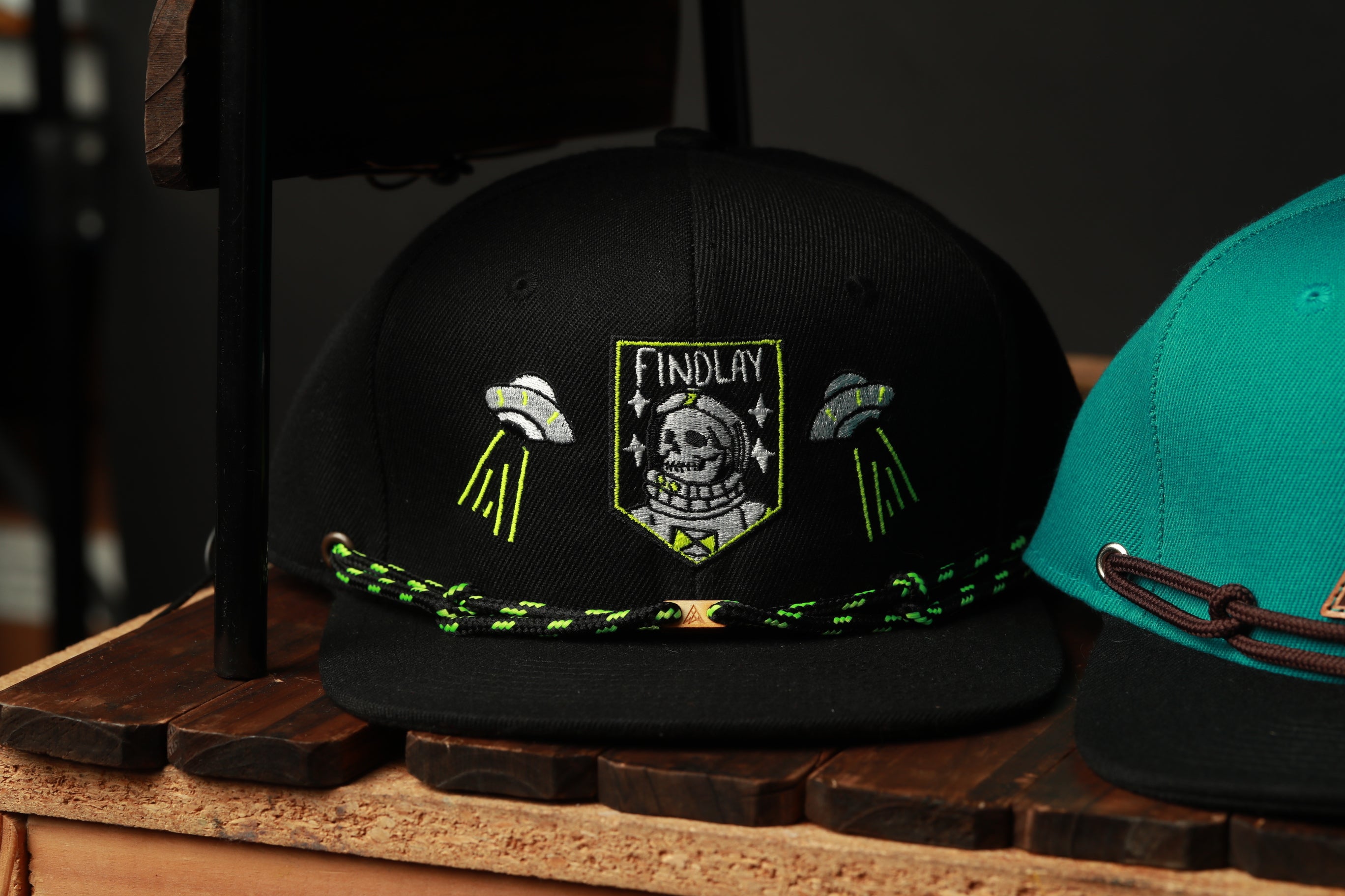 A black baseball cap with a skull and crossbones design, along with the text "FINDLAY" and two green and yellow lines, is displayed on a wooden surface.
