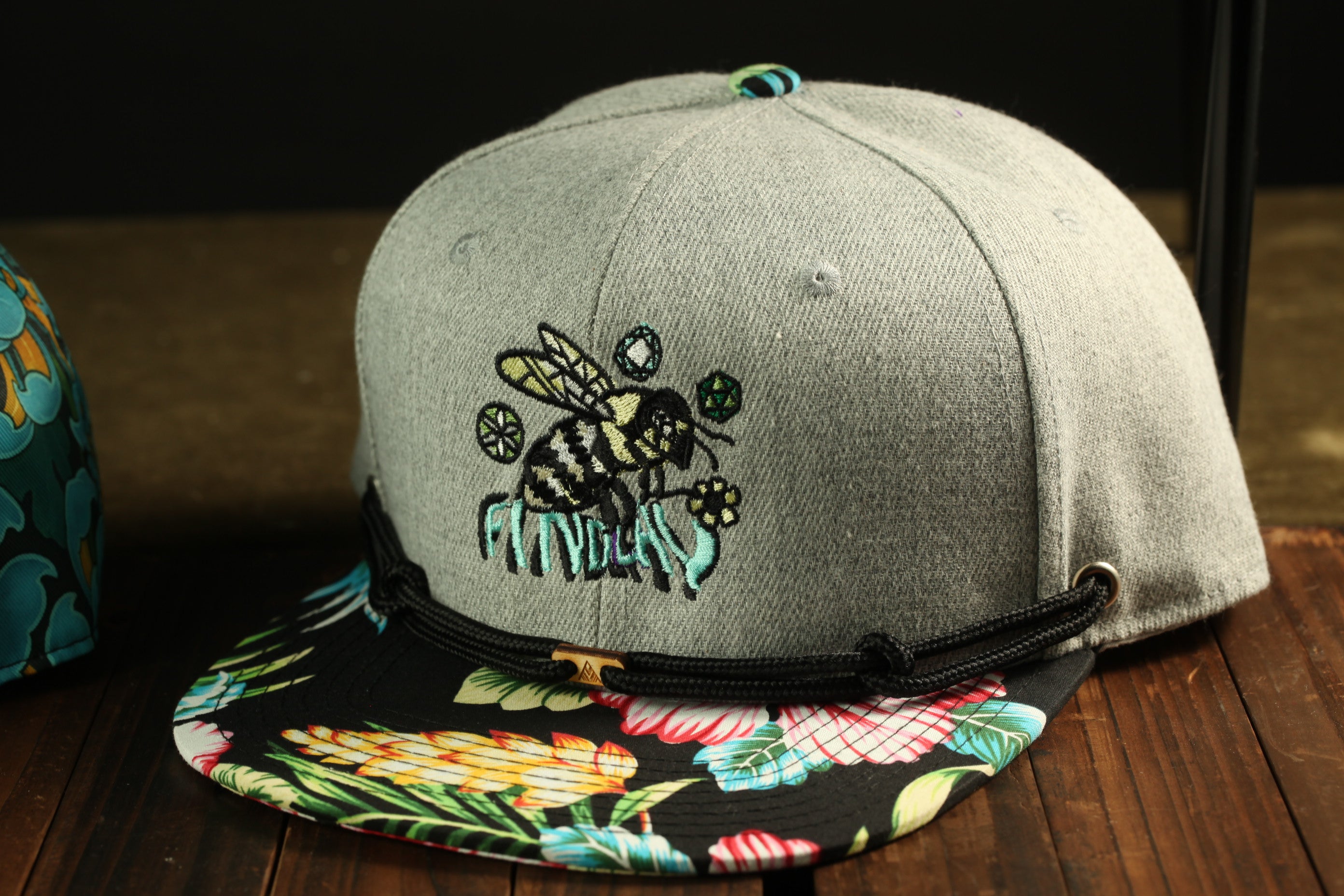 A gray baseball cap with a floral pattern and a bee design, placed on a wooden surface.