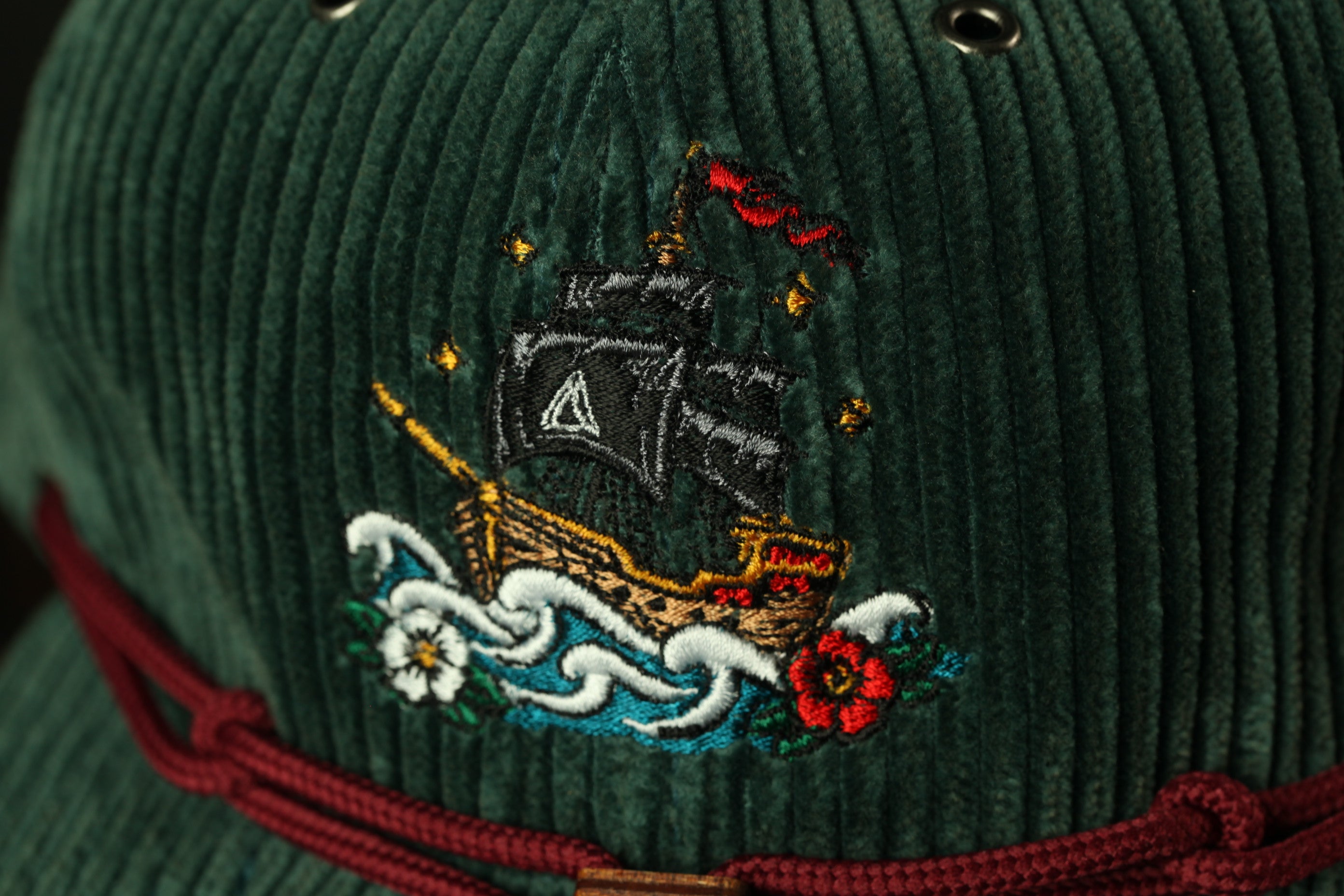 a close-up view of a green corduroy hat with a detailed embroidered design of a ship on the front.