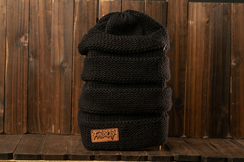 A black knit beanie with a label on it, placed on a wooden surface against a wooden background.