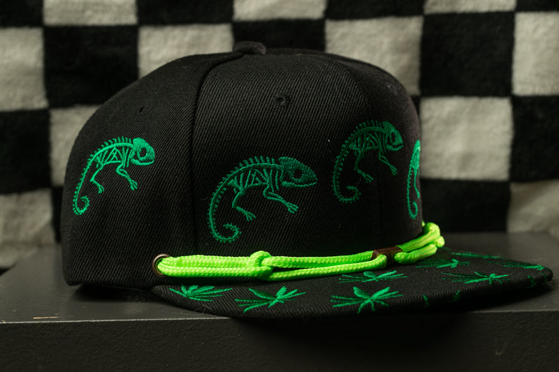 A black baseball cap with green chameleon designs on it, sitting on a black surface against a checkered background.