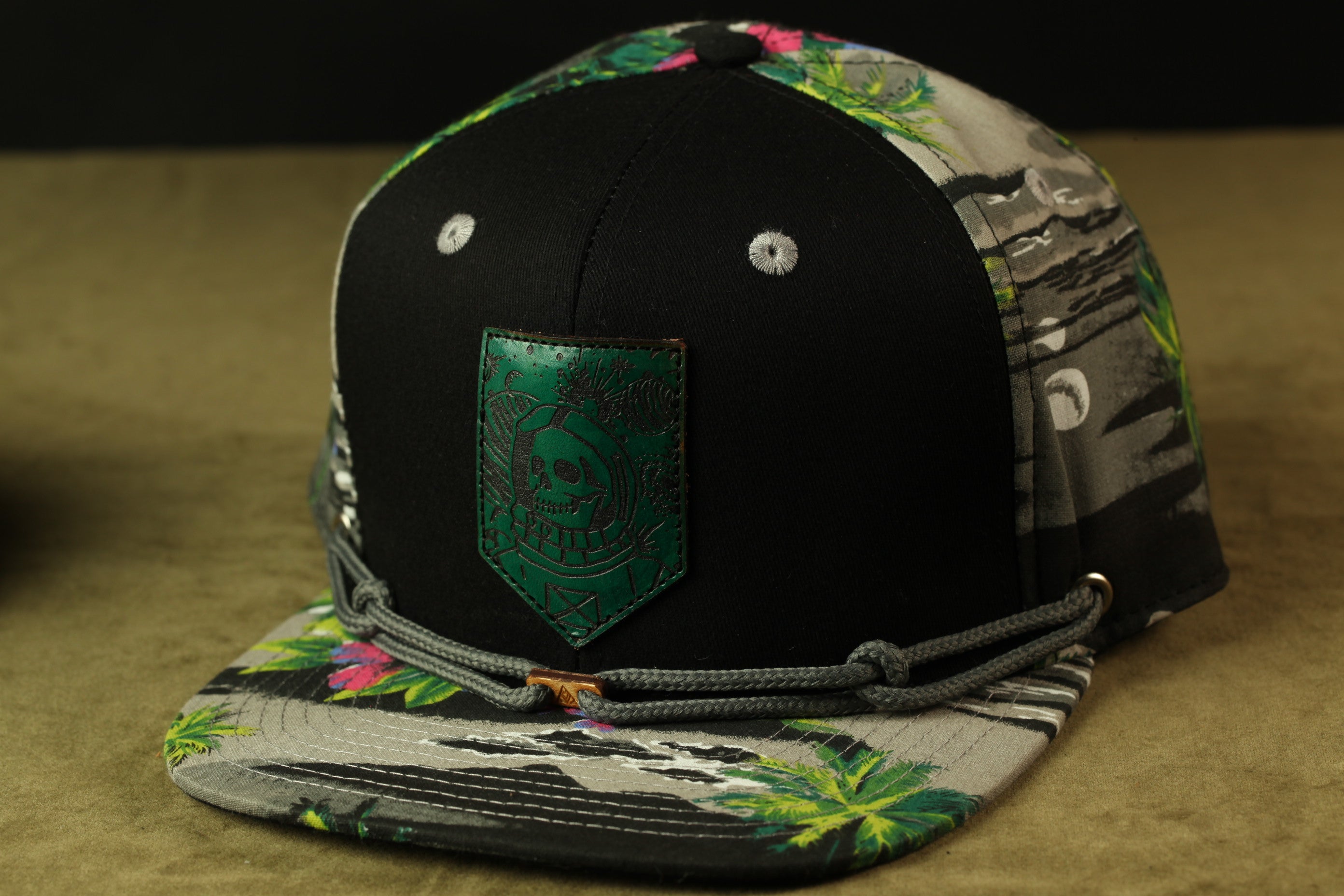 A black baseball cap with a green skull patch and a floral pattern on the brim, sitting on a beige surface.