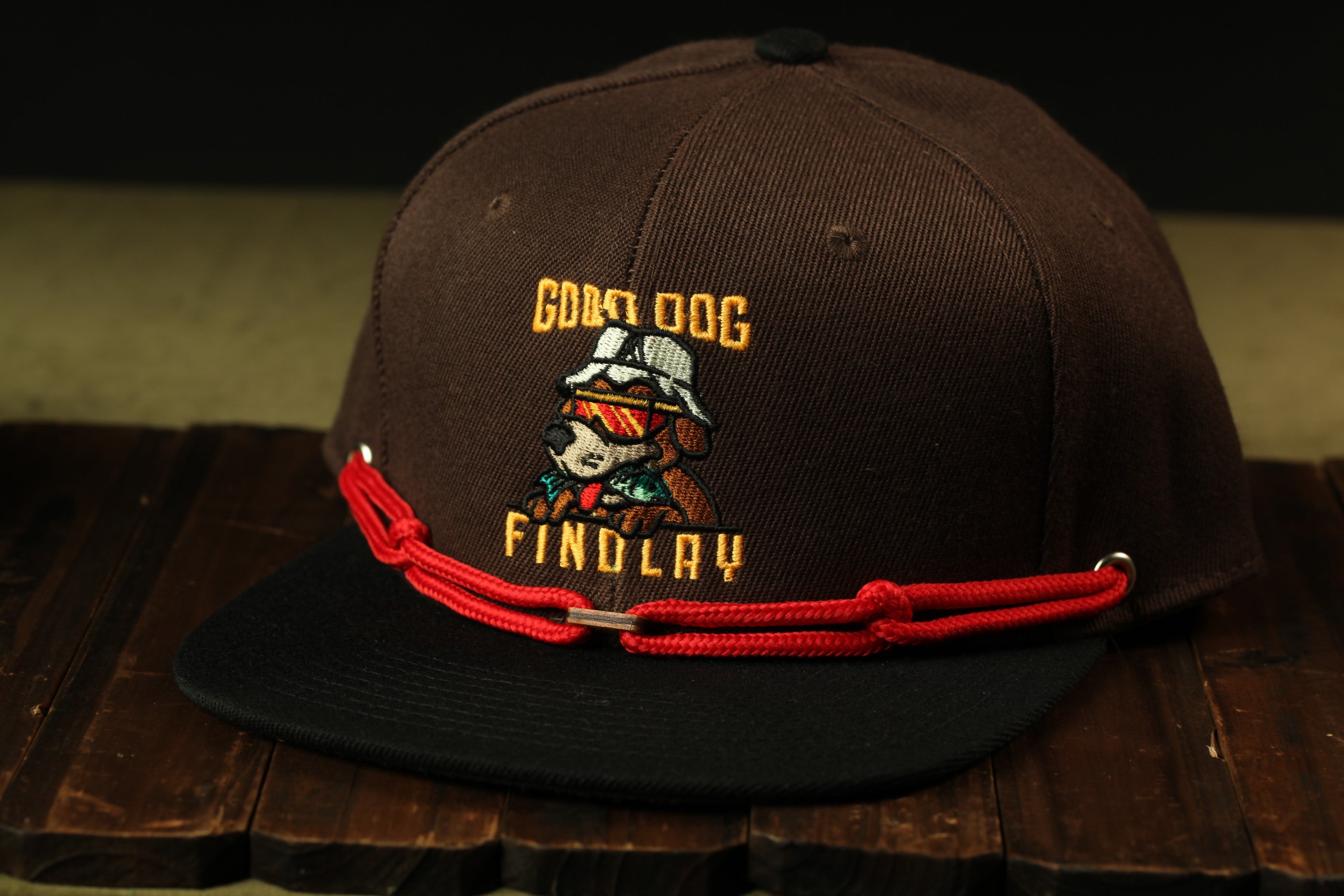 A brown baseball cap with a red string and a cartoon dog logo on it.