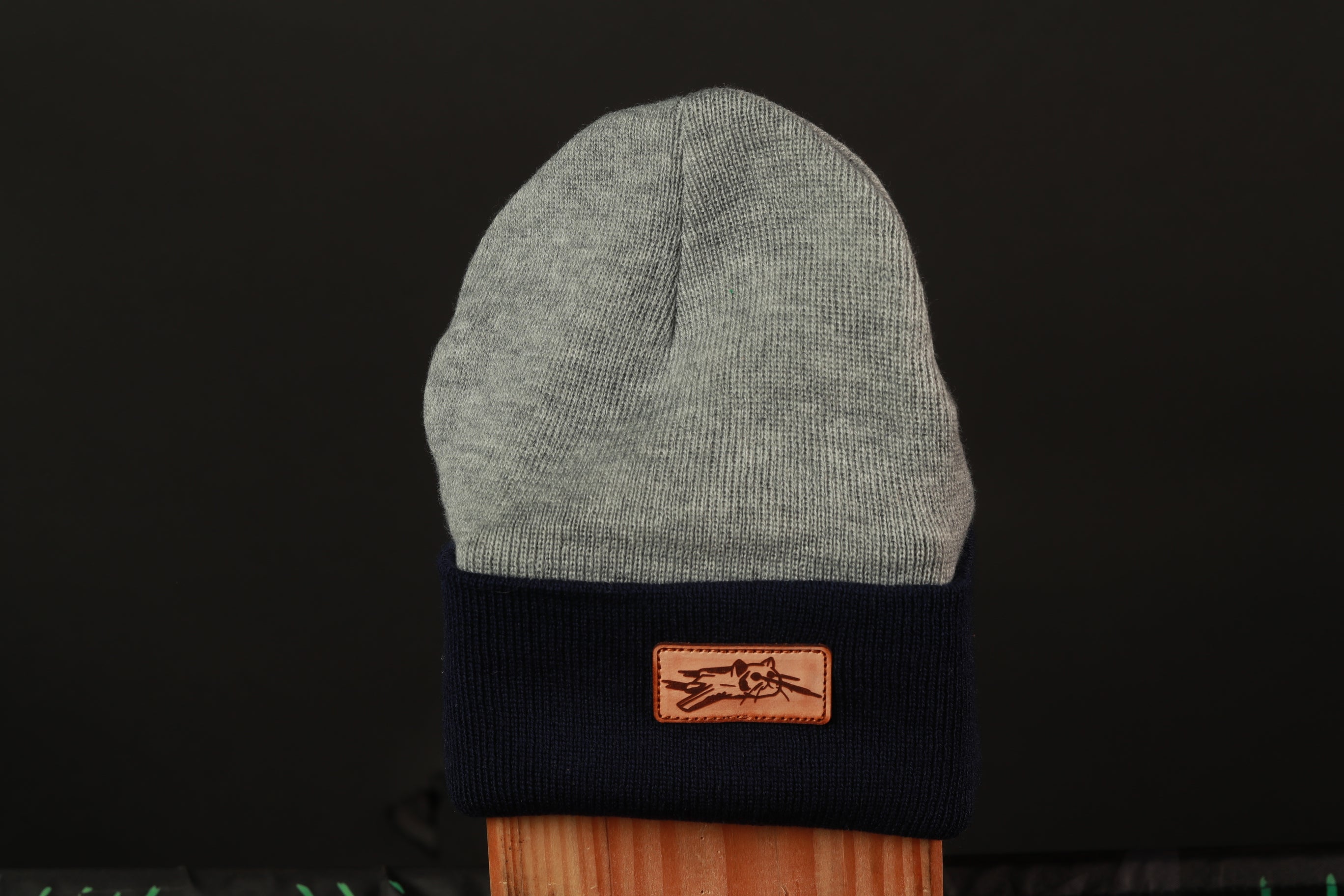 A gray and navy blue knit beanie with a brown leather patch on the front, against a black background.