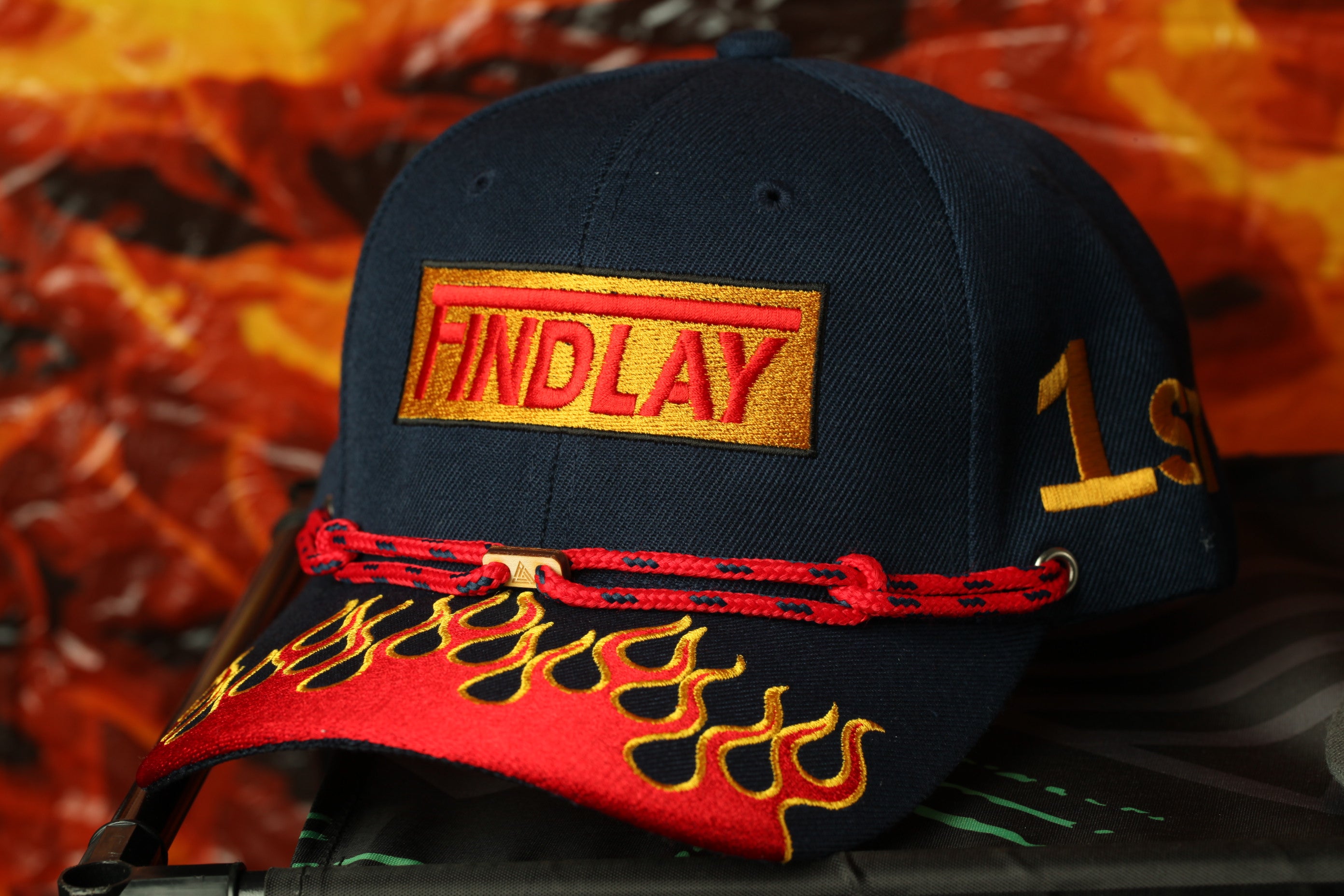 A navy blue baseball cap with a red and gold flame design and the word "FIND LAY" printed on it.