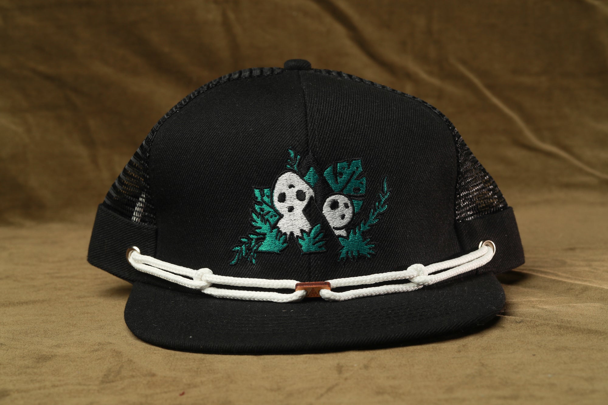 A black baseball cap with a green and white embroidered design featuring two ghostly figures and leaves.