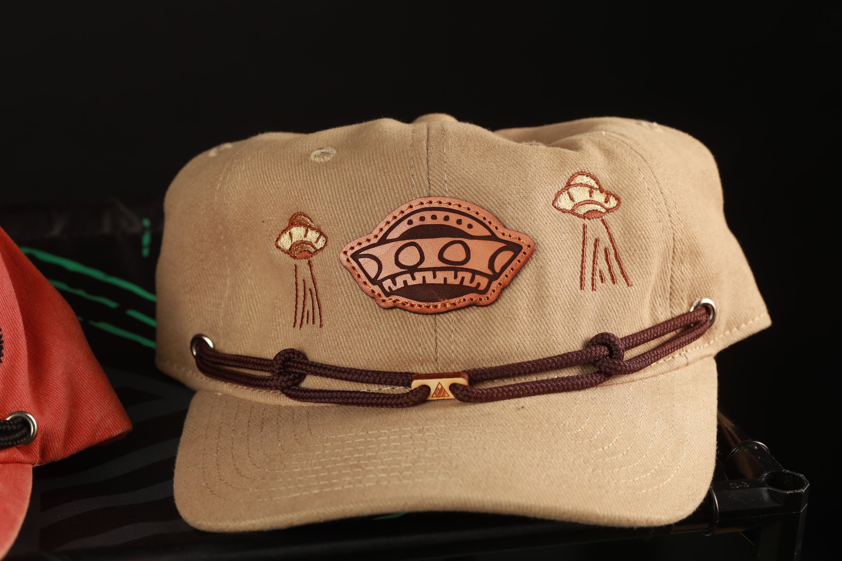 A beige baseball cap with a brown leather strap and a unique alien-like design on the front.