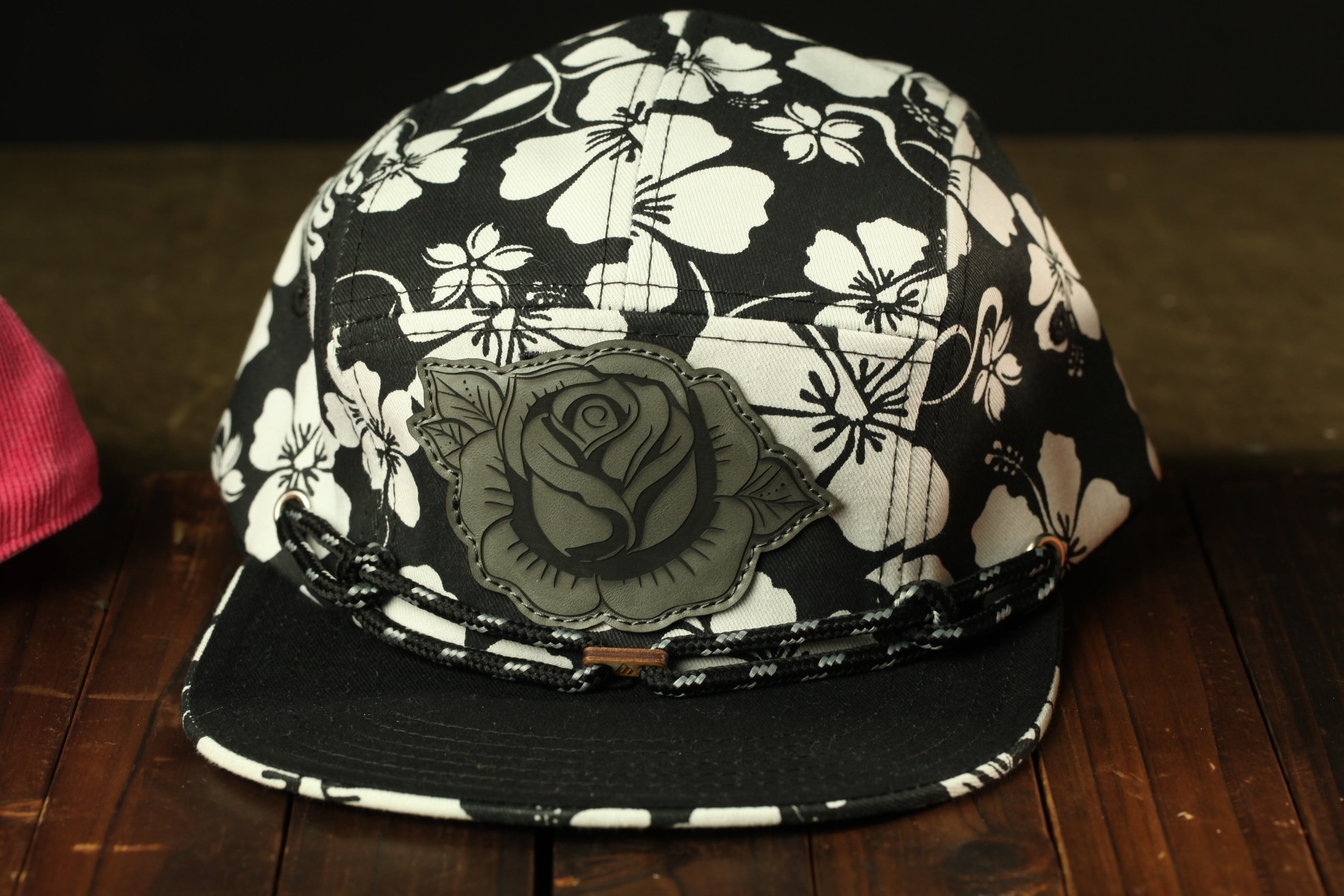 A black and white floral patterned baseball cap with a rose design on the front, resting on a wooden surface.