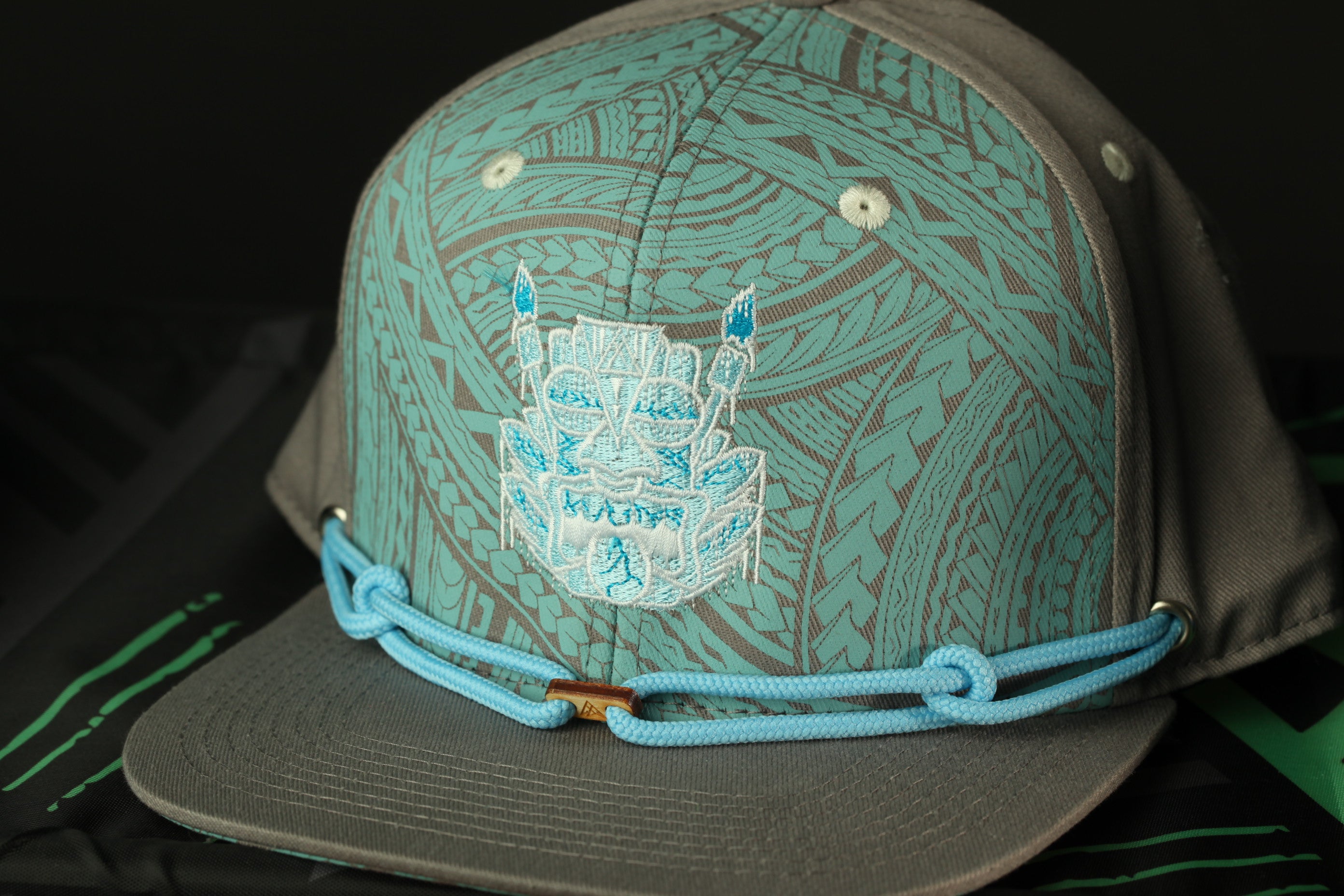 A close-up image of a gray baseball cap with a blue and white patterned design, featuring a central image of a face or mask.