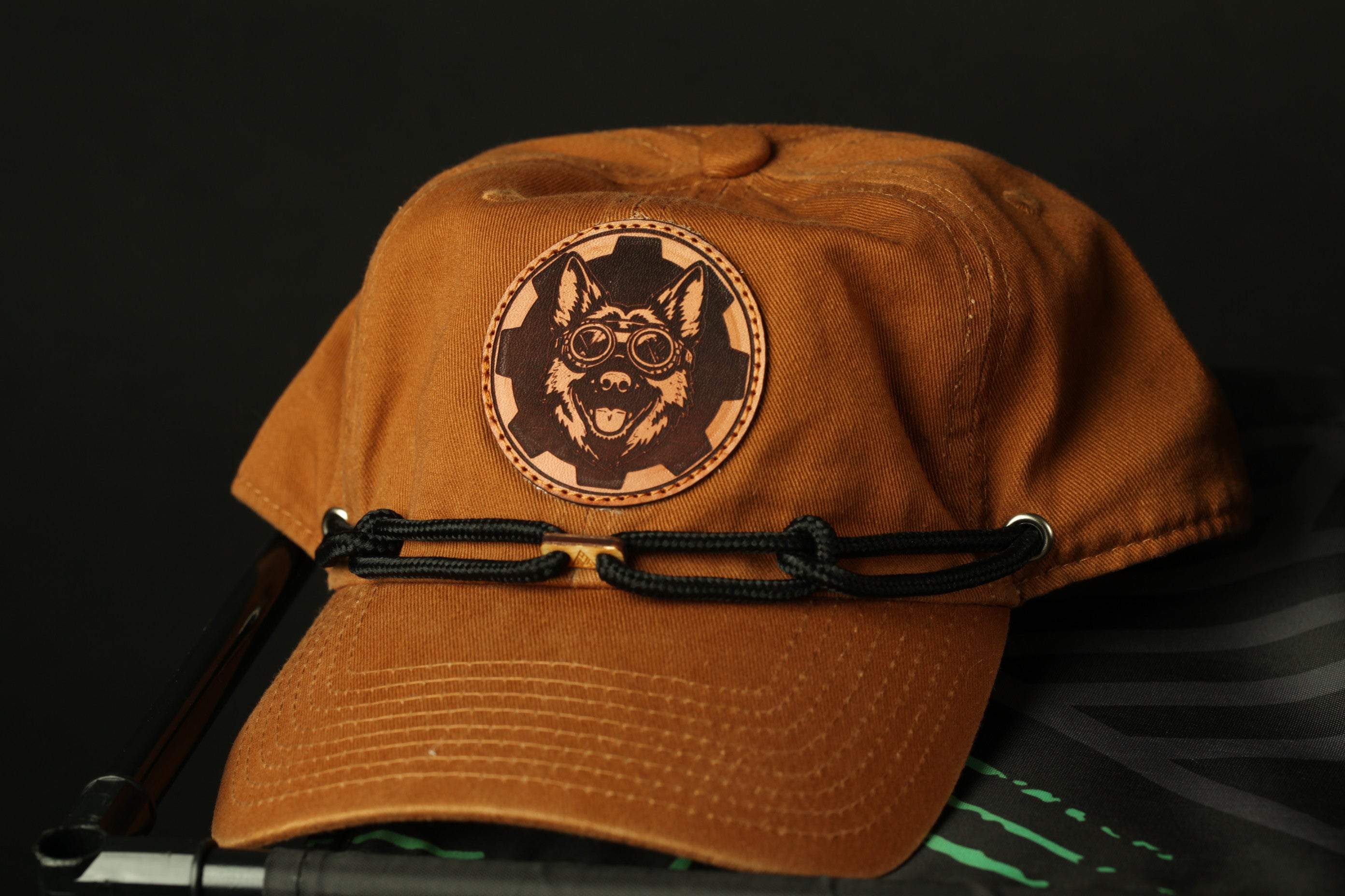A brown baseball cap with a circular patch featuring a dog's face and goggles, along with a black strap, is displayed against a dark background.
