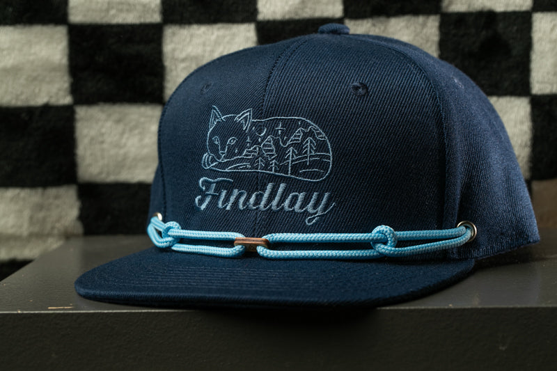 A navy blue baseball cap with a cat design and the word "Findlay" printed on it, sitting on a black surface against a checkered background.