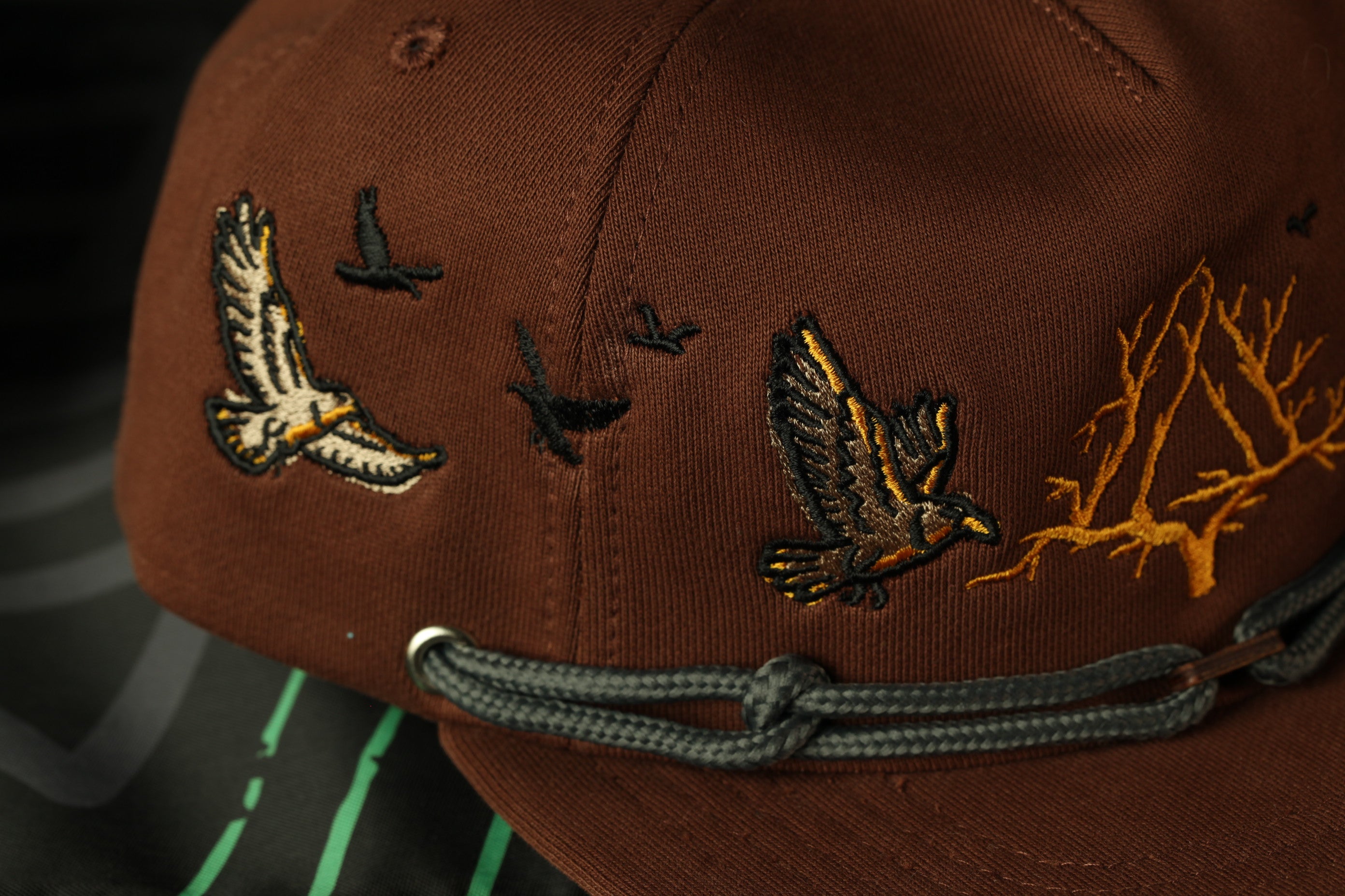 A close-up view of a brown baseball cap with embroidered designs of birds and trees on the front.