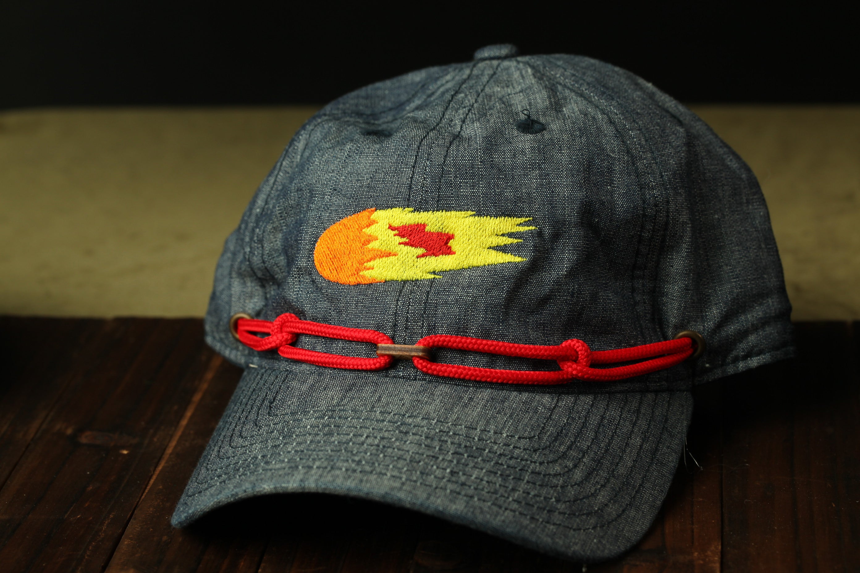 A denim baseball cap with a red string and a yellow and orange flame design on the front.