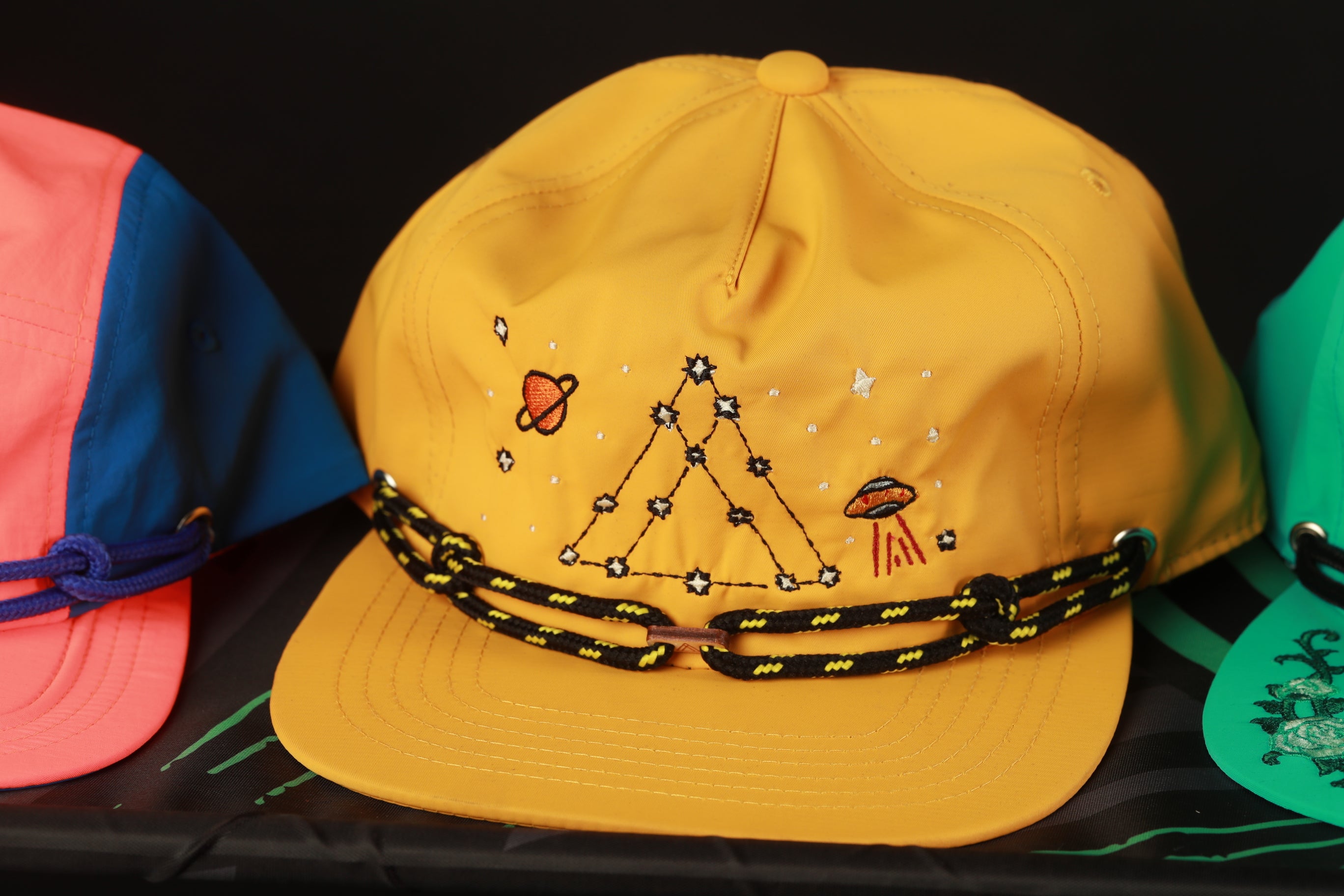 A yellow baseball cap with a black and yellow string, featuring an embroidered design of stars and a planet, is displayed on a table.