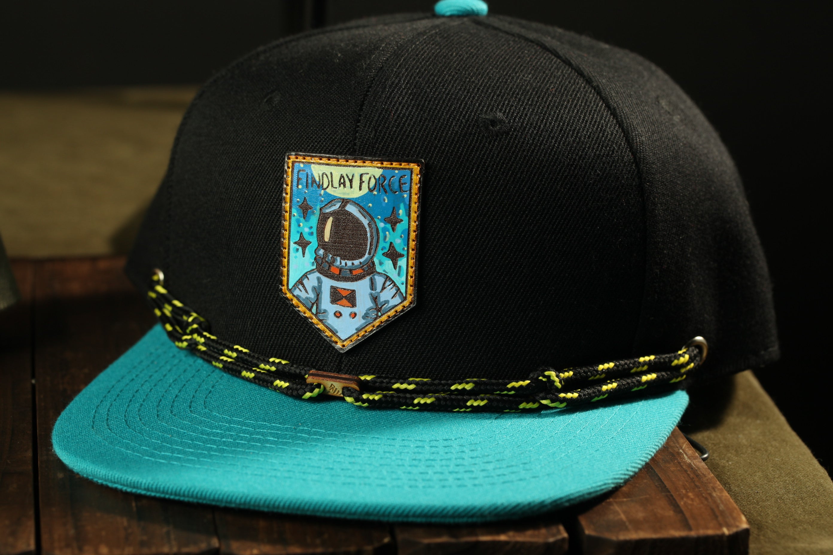 A black baseball cap with a teal brim and a patch featuring an astronaut, along with a black and yellow string around the brim.