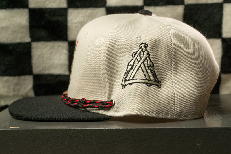 A white baseball cap with a black brim and a red and black braided strap is shown against a black and white checkered background.