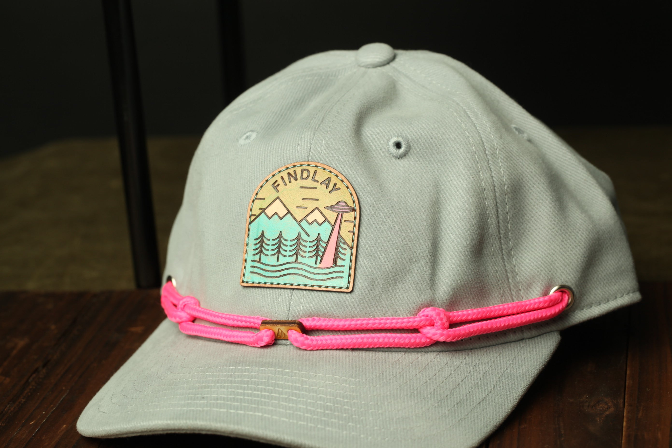 A gray baseball cap with a pink braided strap and a patch on the front, featuring a mountain landscape and an alien.