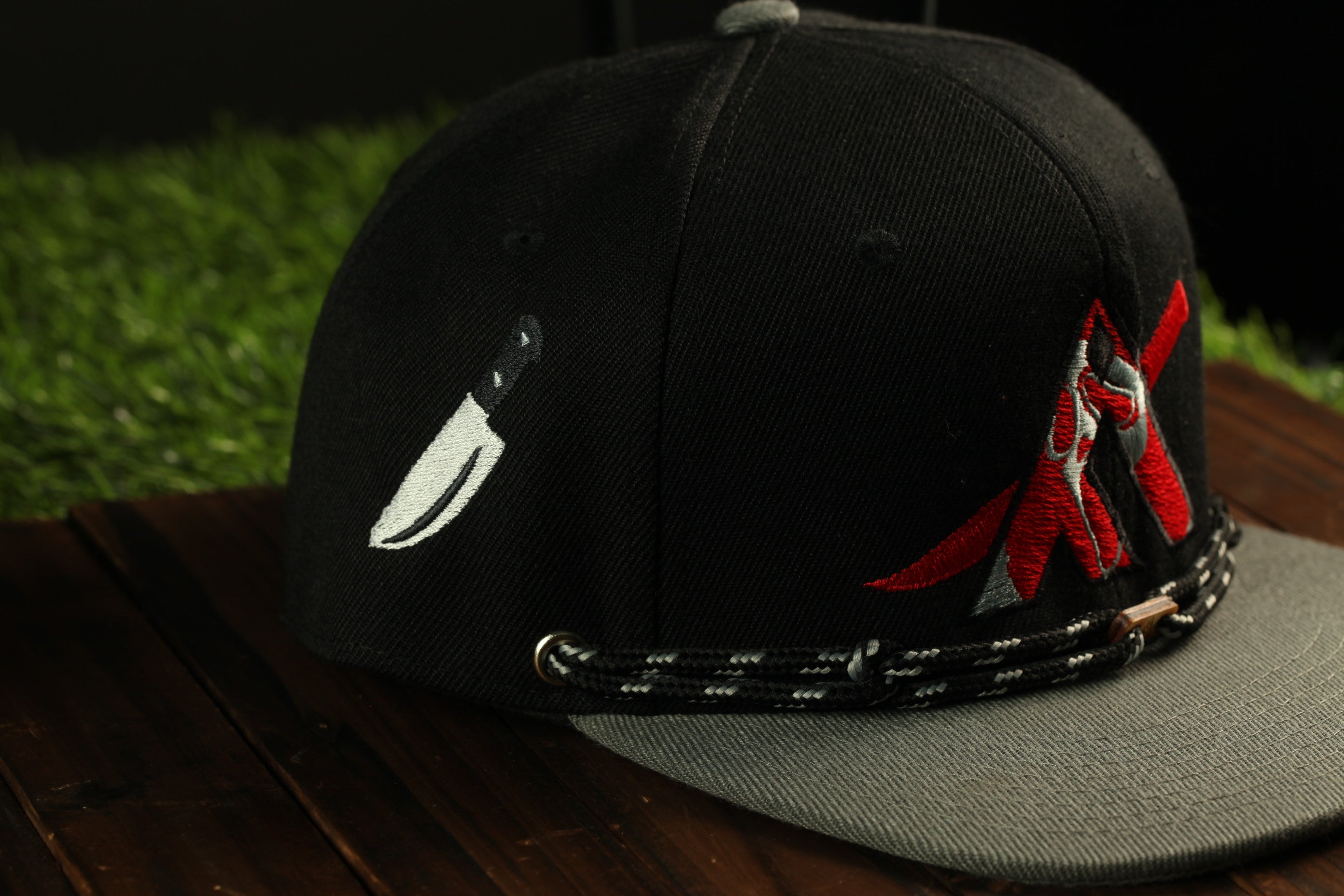 A black baseball cap with a knife embroidered on it, placed on a wooden surface.