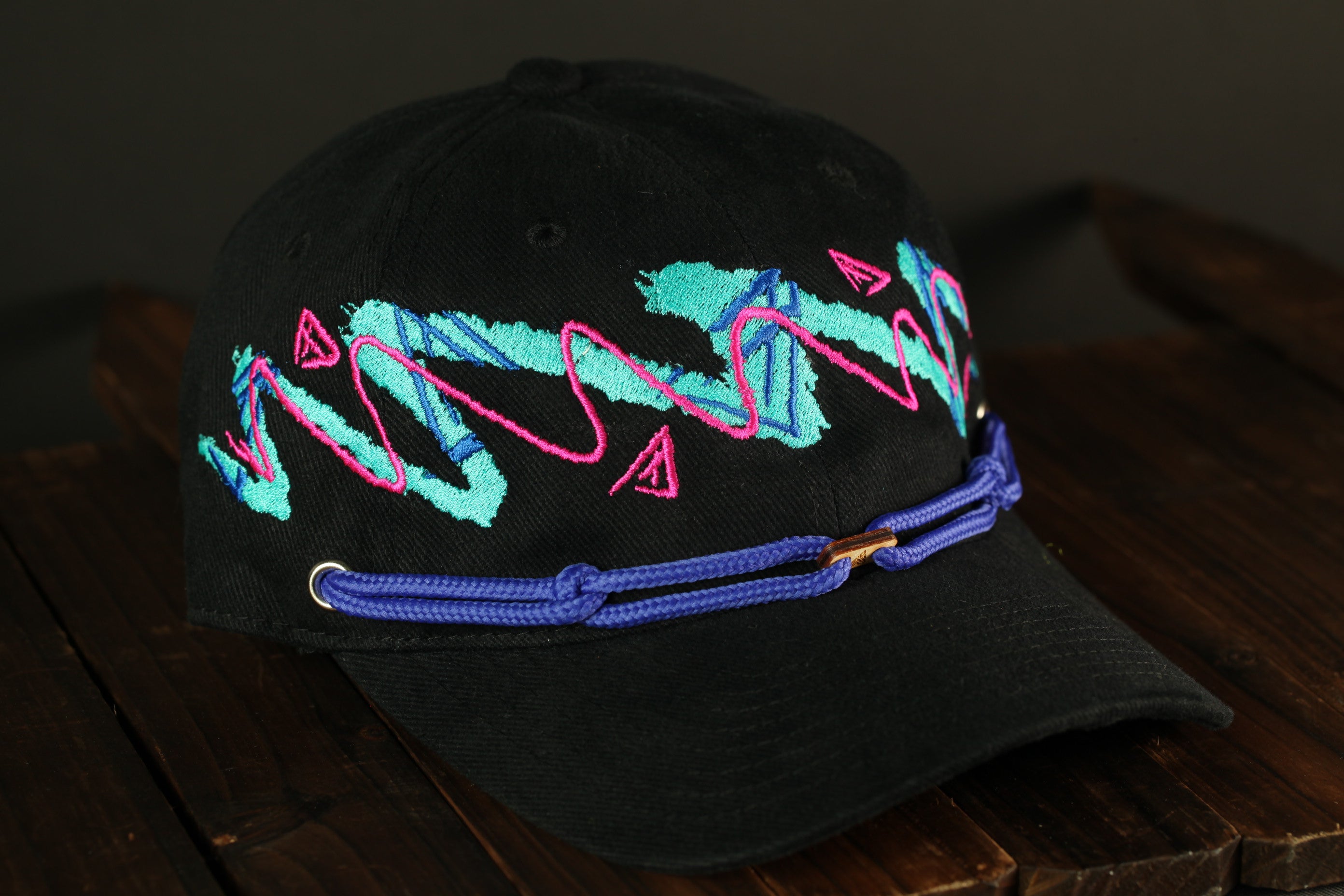 A black baseball cap with colorful embroidered designs on the front, including a pink and blue wave pattern and other abstract shapes.