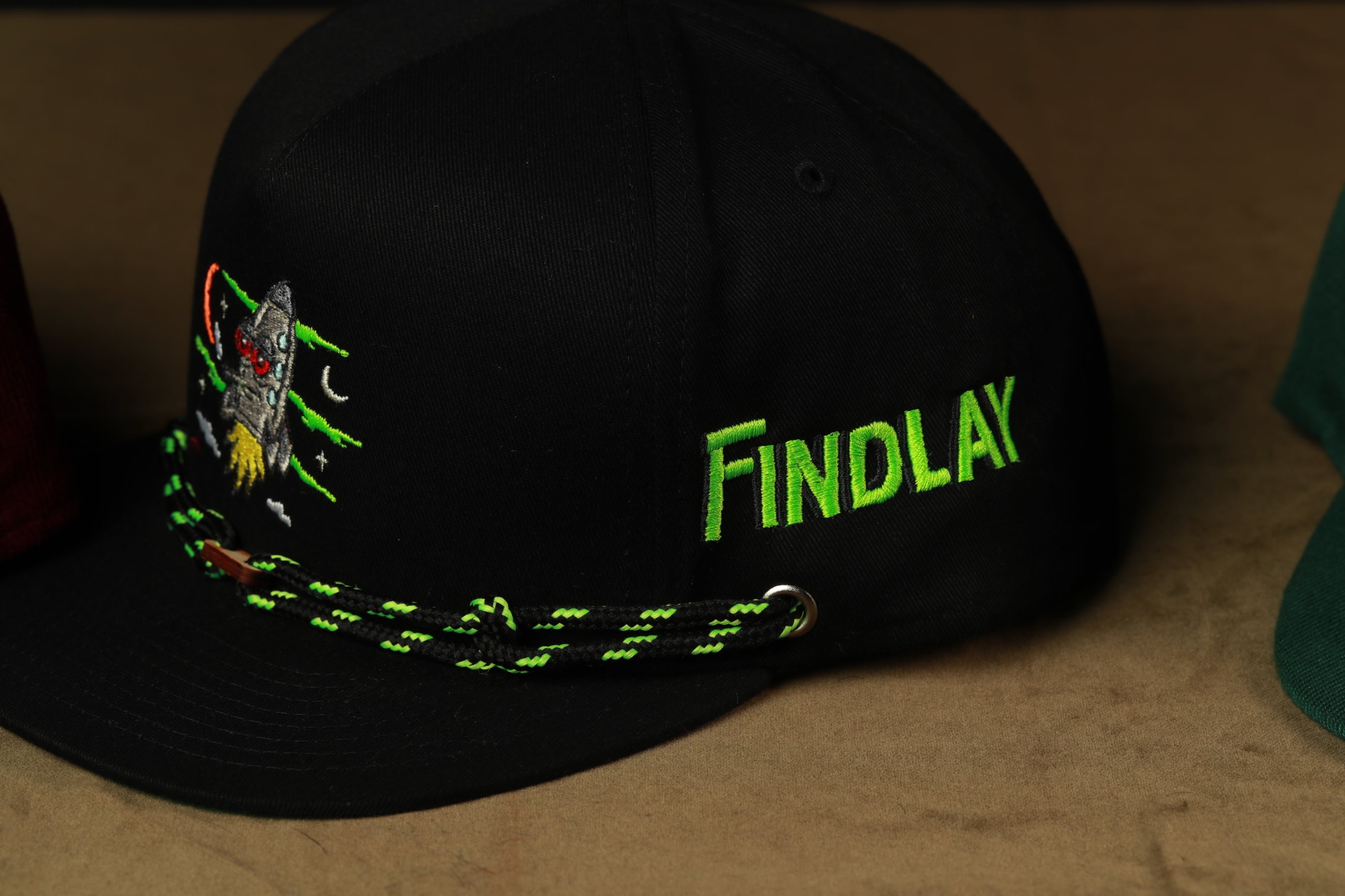 A black baseball cap with a green and black braided strap, featuring a small embroidered design and the word "FINDLAY" printed on the side.