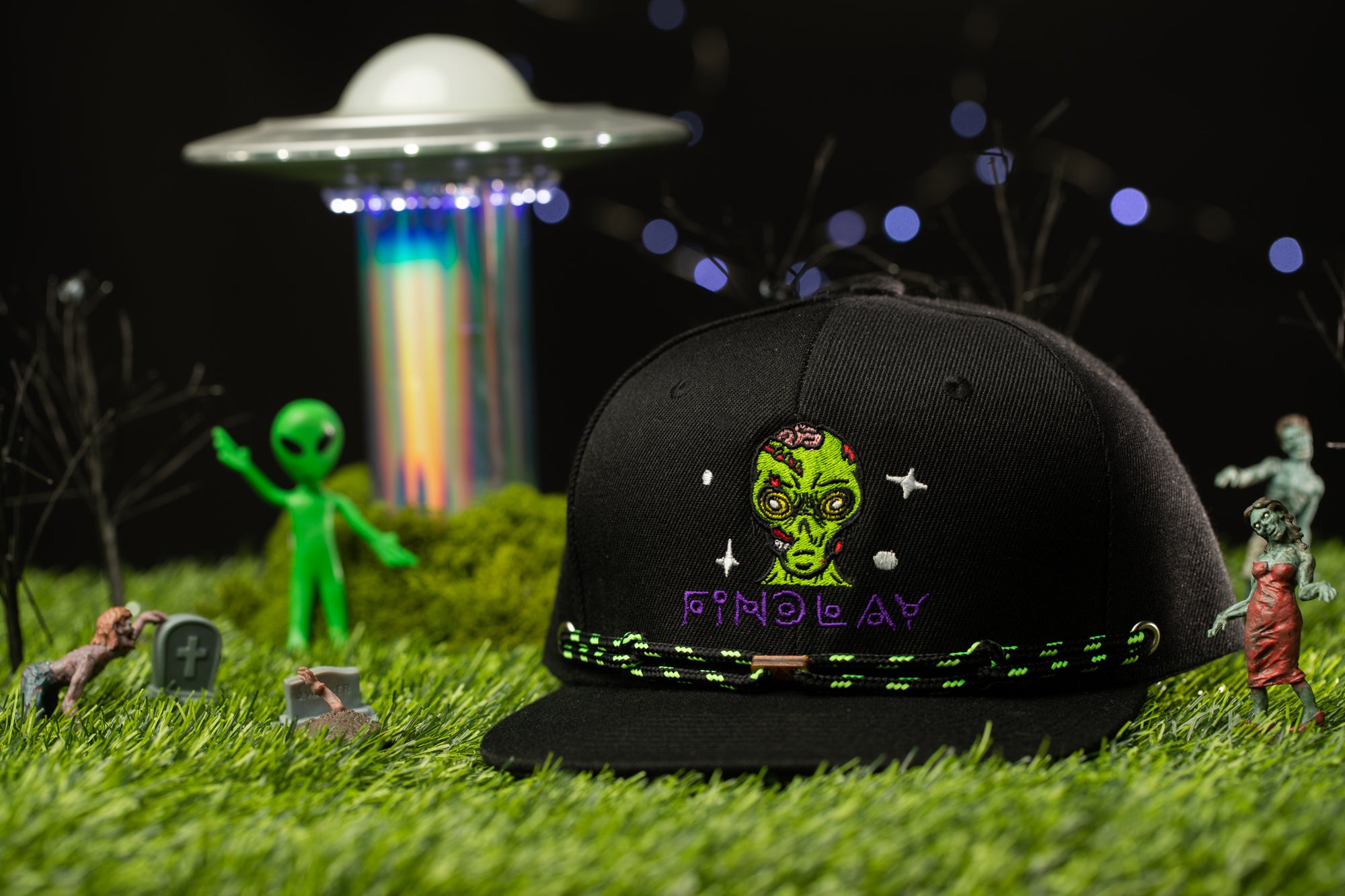 a black baseball cap with a green alien face and the word "Finlay" printed on it, surrounded by various small toy figures and a green alien figure in a grassy field.