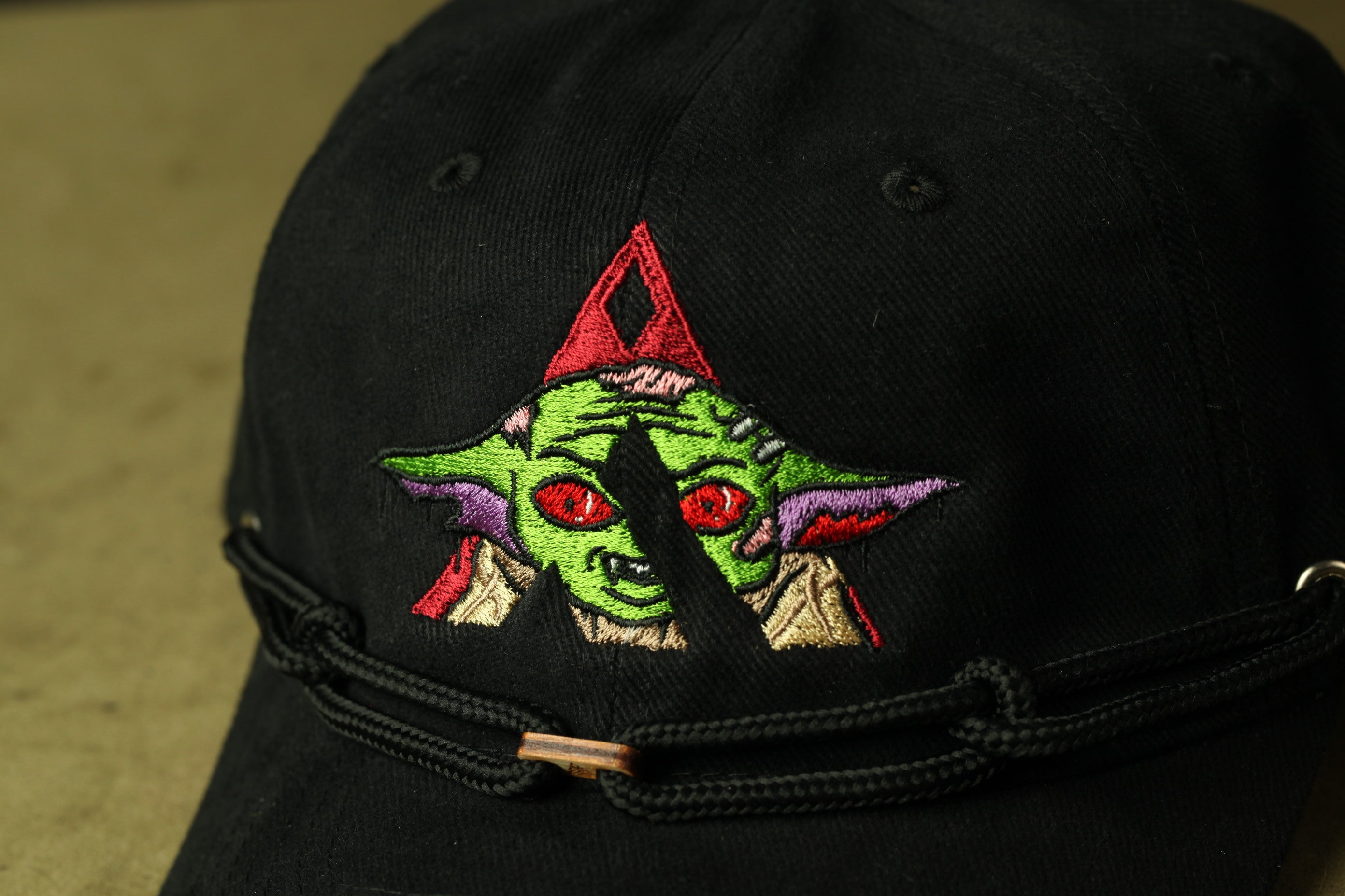 A close-up view of a black baseball cap with a colorful, cartoonish design featuring a green alien-like creature with red eyes and a red triangle on the front.