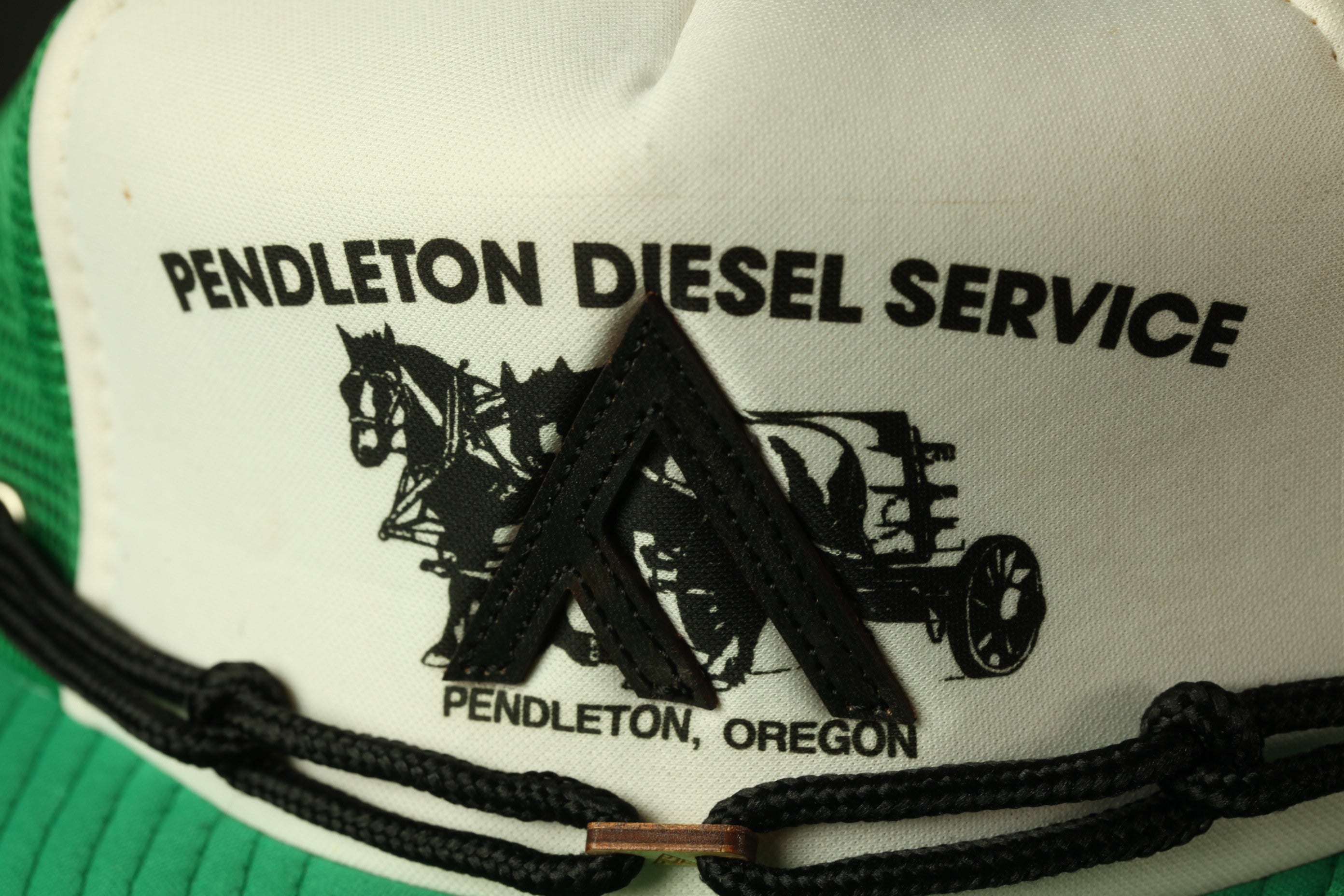 A close-up of a white and green baseball cap with the text "PENDLETON DIESEL SERVICE" and an illustration of a horse-drawn carriage.