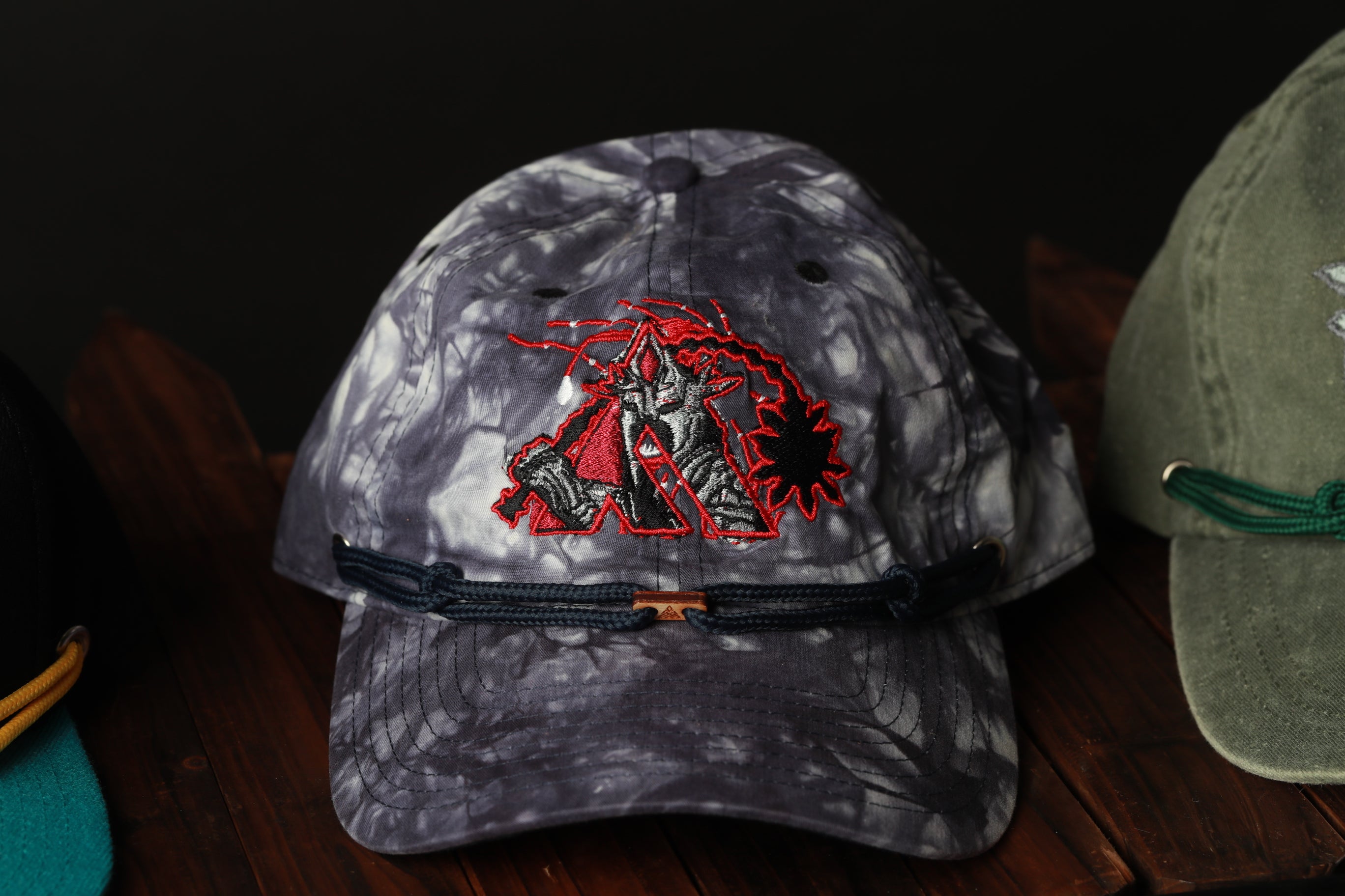 A gray and red tie-dye baseball cap with a dragon embroidered on it, displayed on a wooden surface.