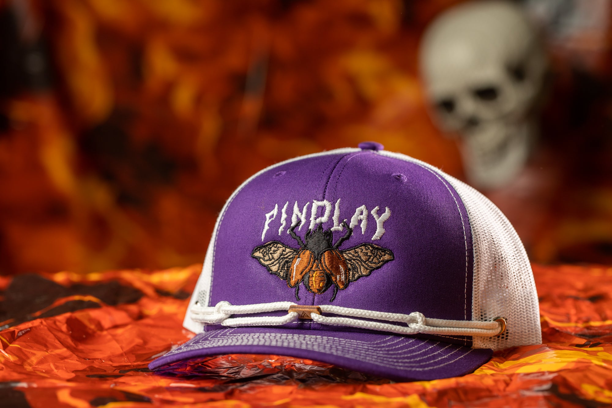 A purple baseball cap with a white mesh panel and a graphic of a fly is displayed against a background of orange and red leaves.