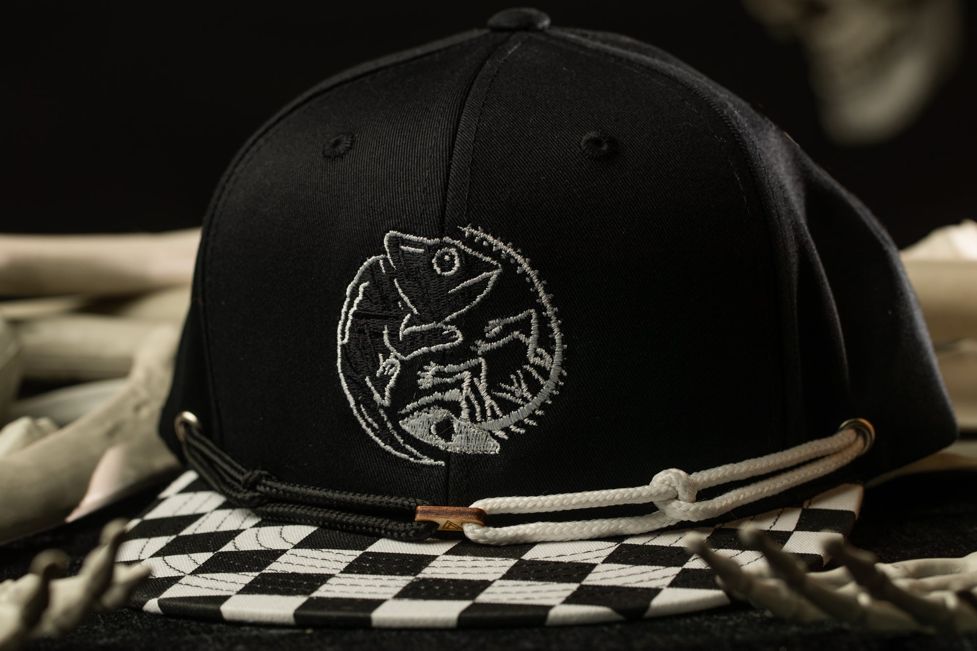 A black baseball cap with a white embroidered design on the front, featuring a chameleon or similar creature.