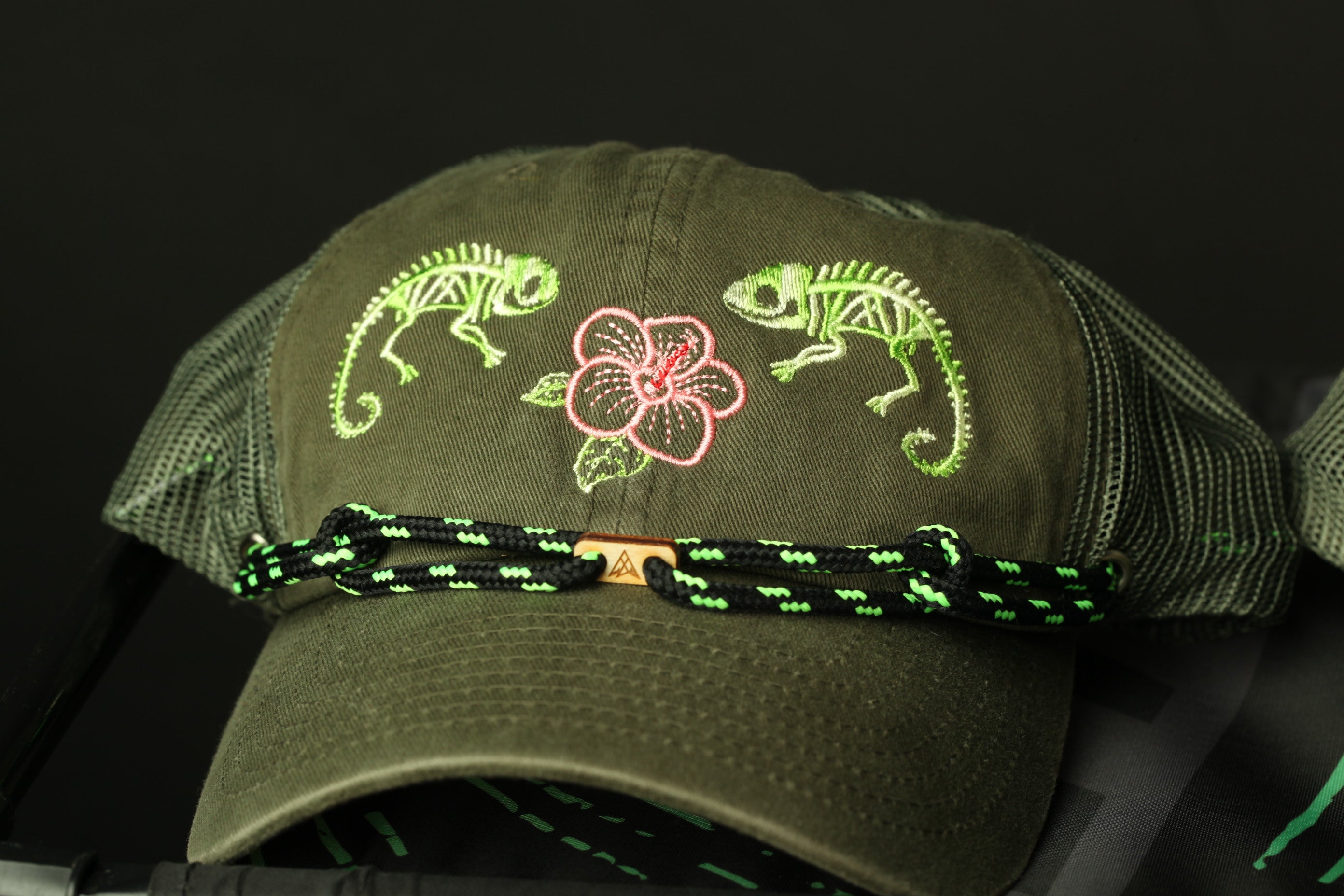 A green baseball cap with a floral design and two green chameleons on it, along with a black and green braided strap.