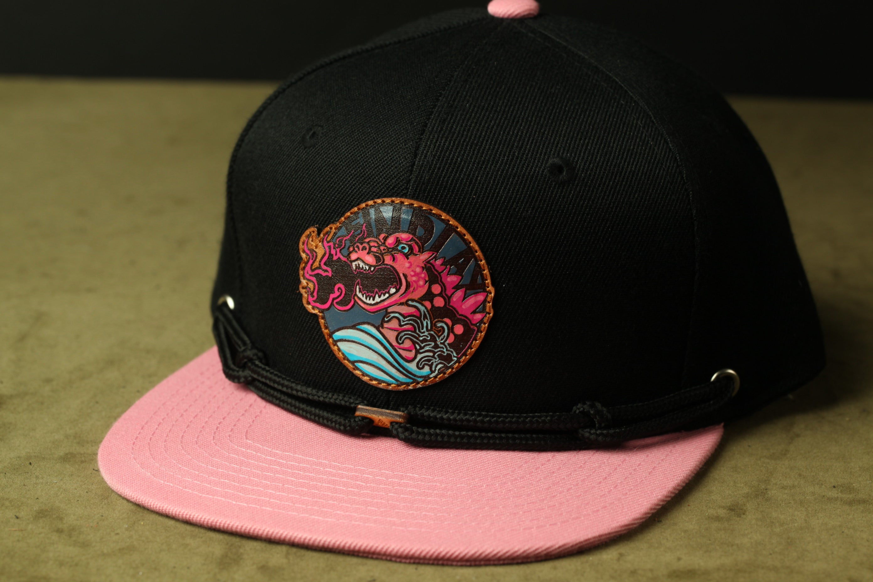A black baseball cap with a pink brim and a colorful, cartoonish dragon design on the front.