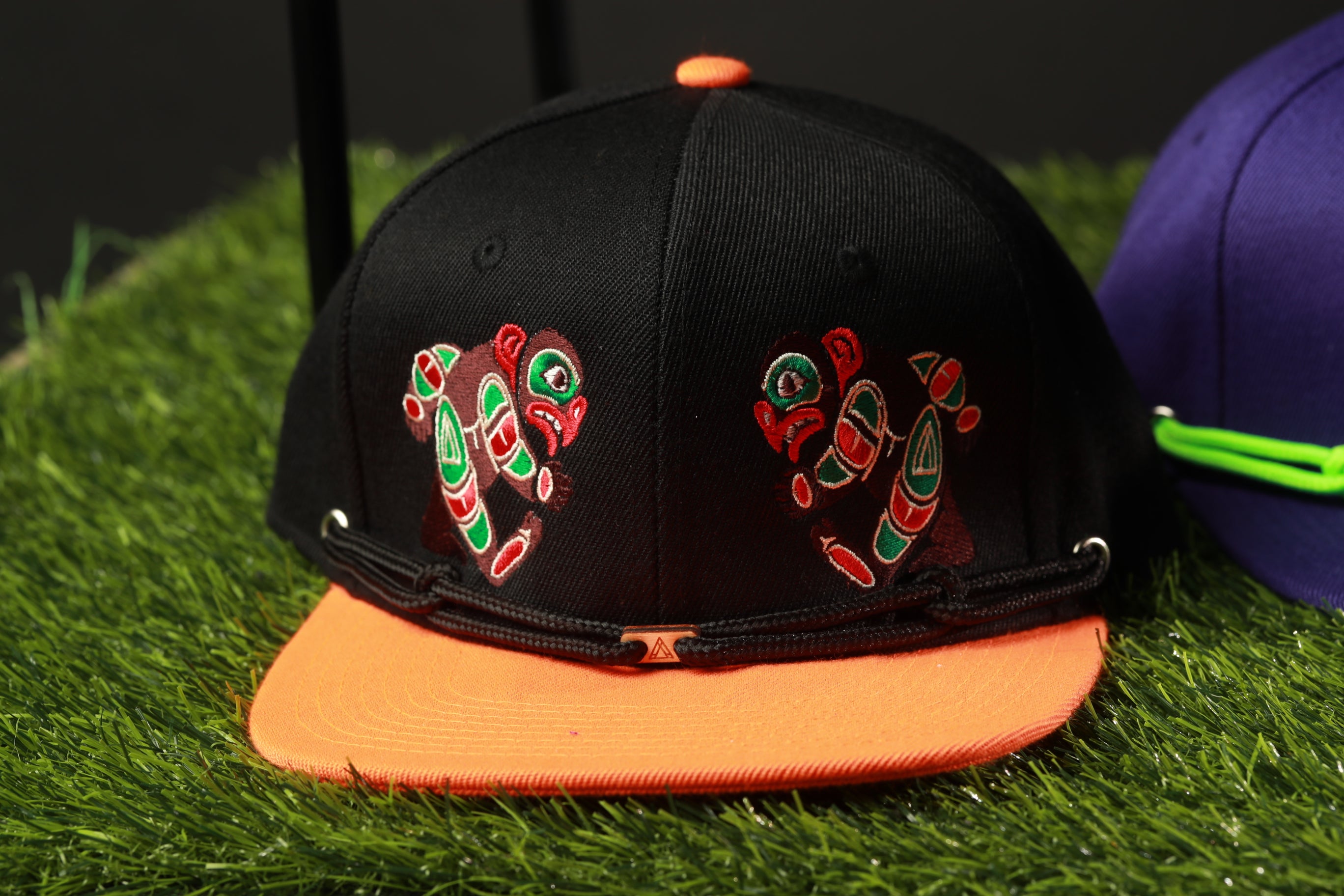 A black baseball cap with colorful designs on it, placed on a grassy surface next to another cap.