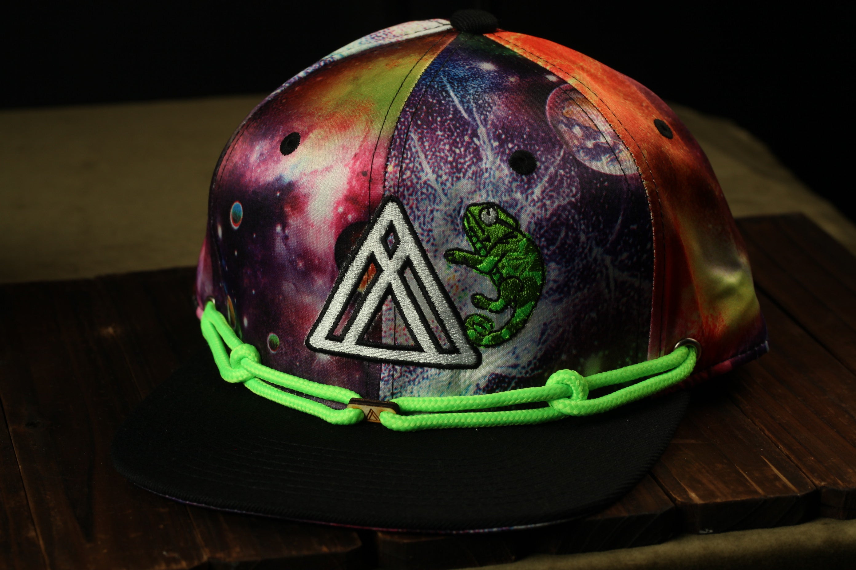 A colorful, patterned baseball cap with a space-themed design, featuring a green chameleon and various celestial elements.