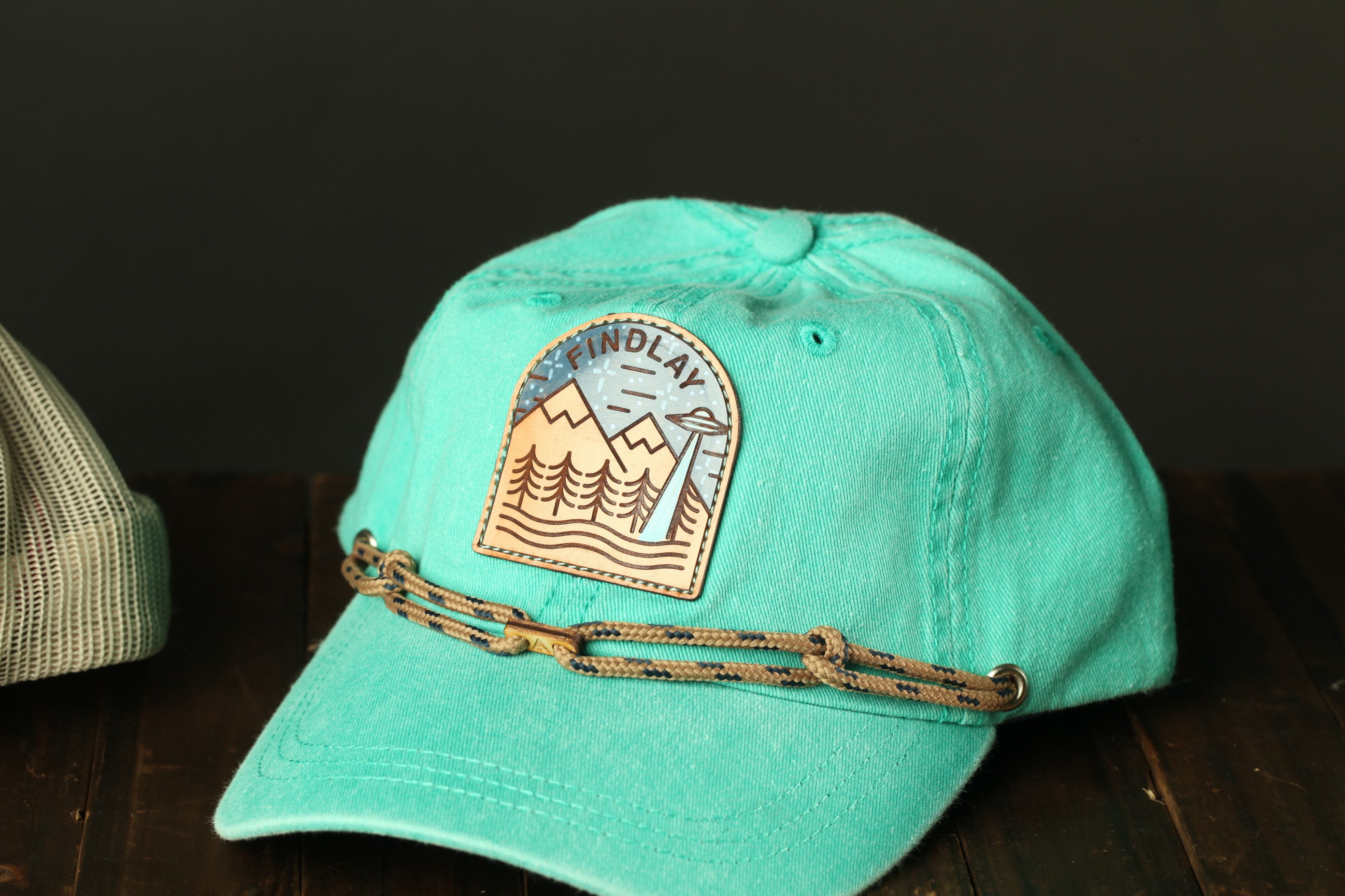 A turquoise baseball cap with a mountain-themed patch and a rope-like strap is displayed on a wooden surface.