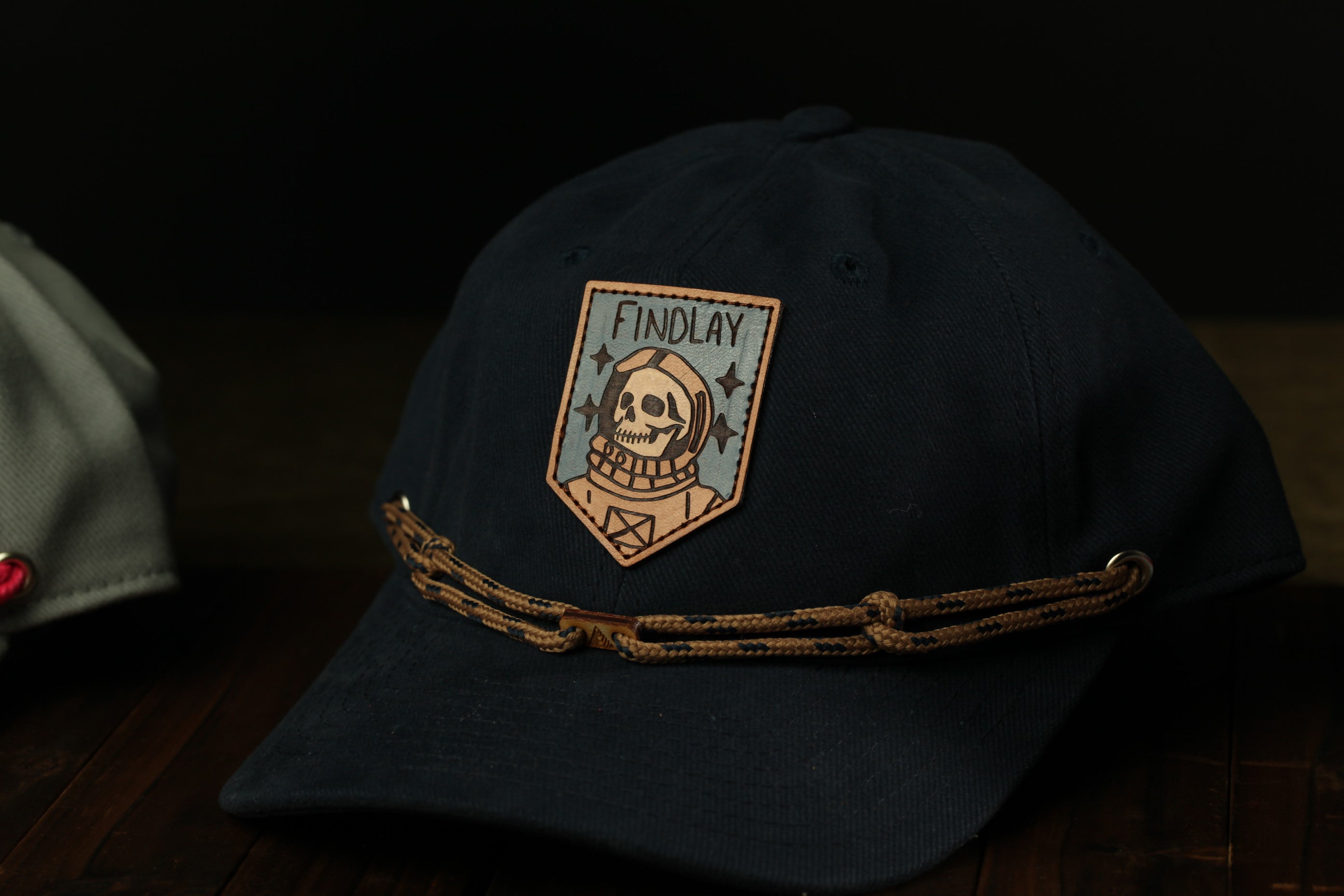 A dark blue baseball cap with a skull patch and the word "FINDOLAY" printed on it, along with a brown chain strap.