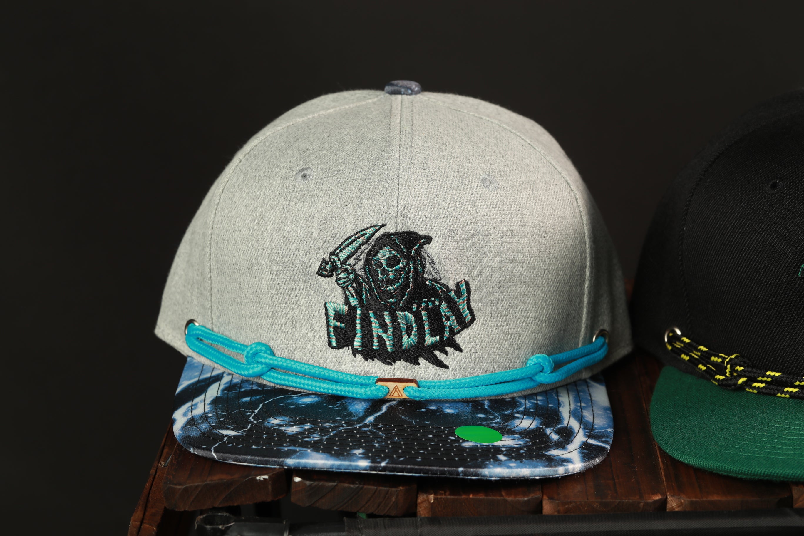 A gray baseball cap with a skull and crossbones design and the word "FINLAND" printed on it, placed on a wooden surface next to another black baseball cap.