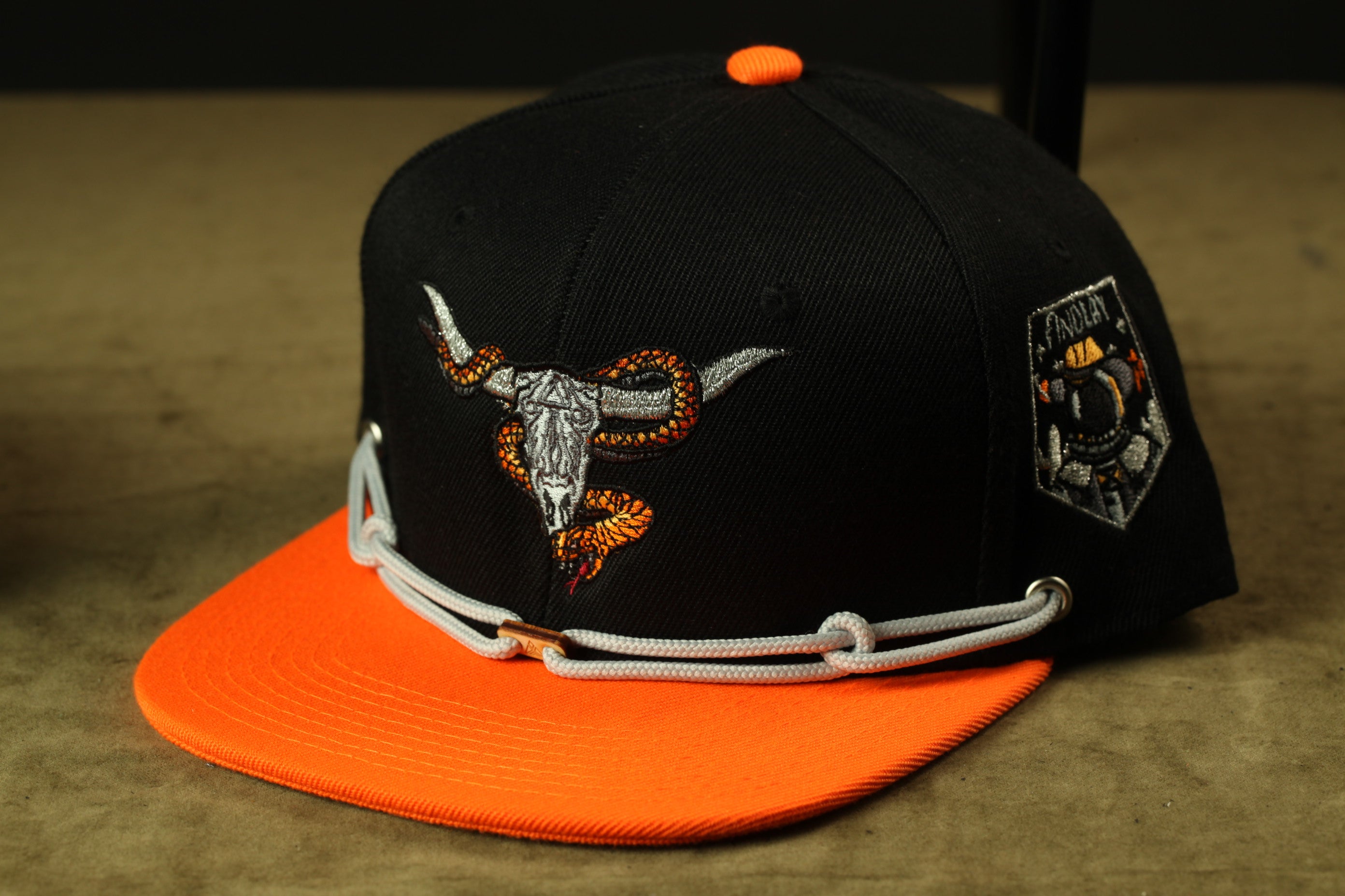 A black baseball cap with an orange brim and a detailed embroidered design of a bull's head and snake on the front.