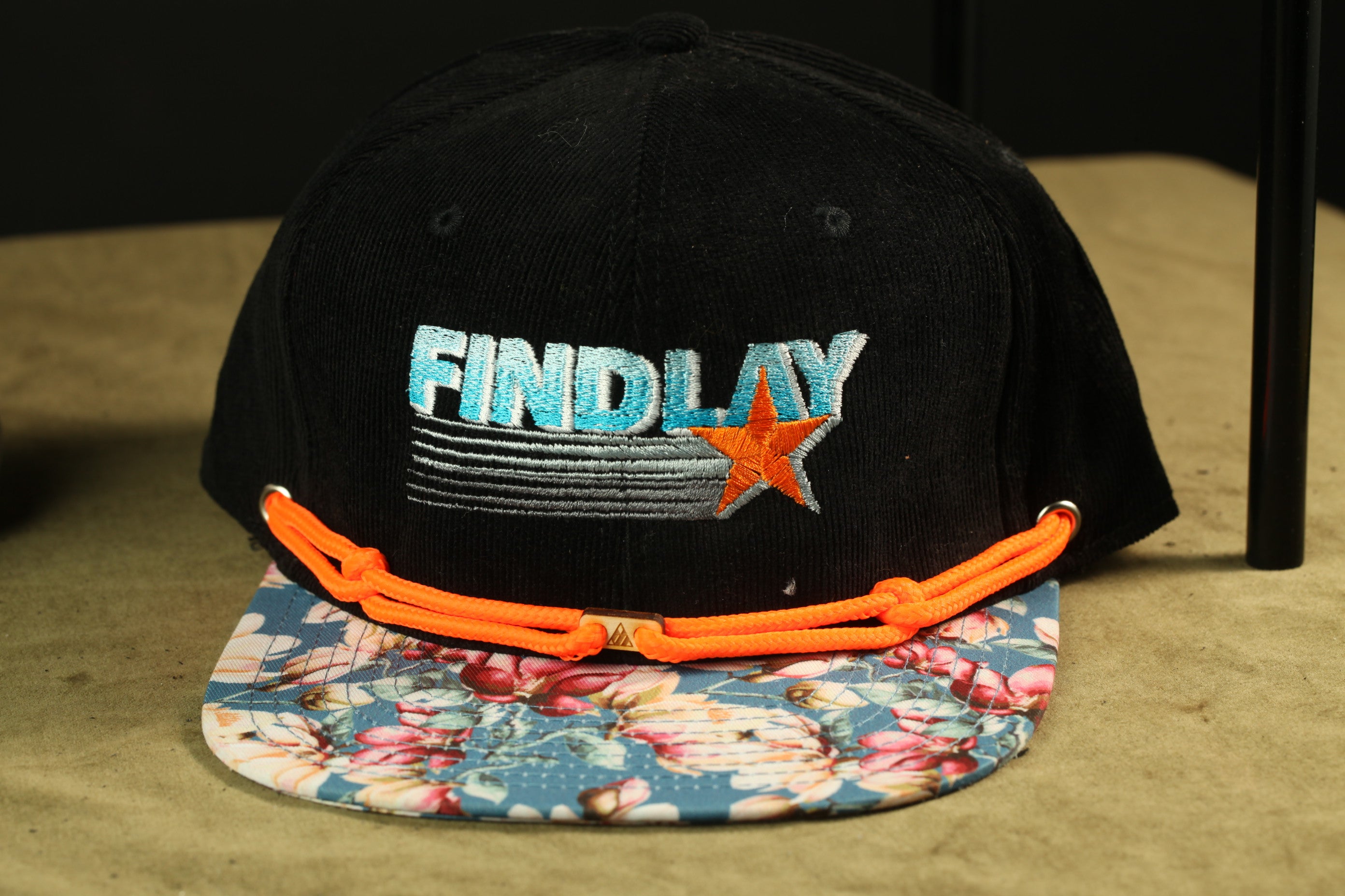 A black baseball cap with a floral pattern and orange straps, featuring the word "FINDlay" embroidered on it.