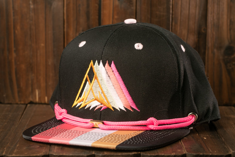 A black baseball cap with a colorful geometric pattern and pink strings is displayed on a wooden surface.