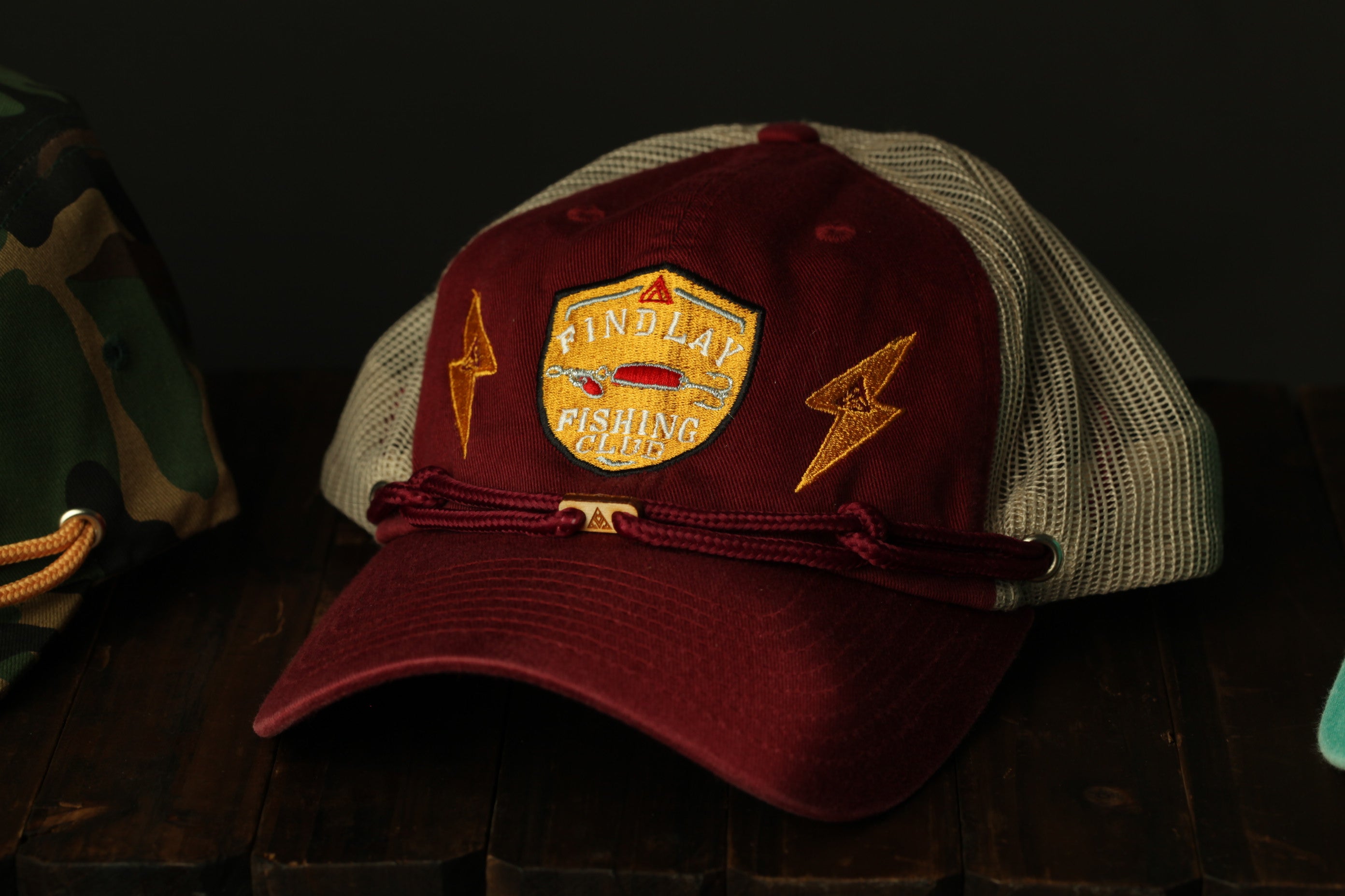 A maroon baseball cap with a mesh back and a gold emblem, displaying the text "FIND LAY FISHING CLUB" on the front.