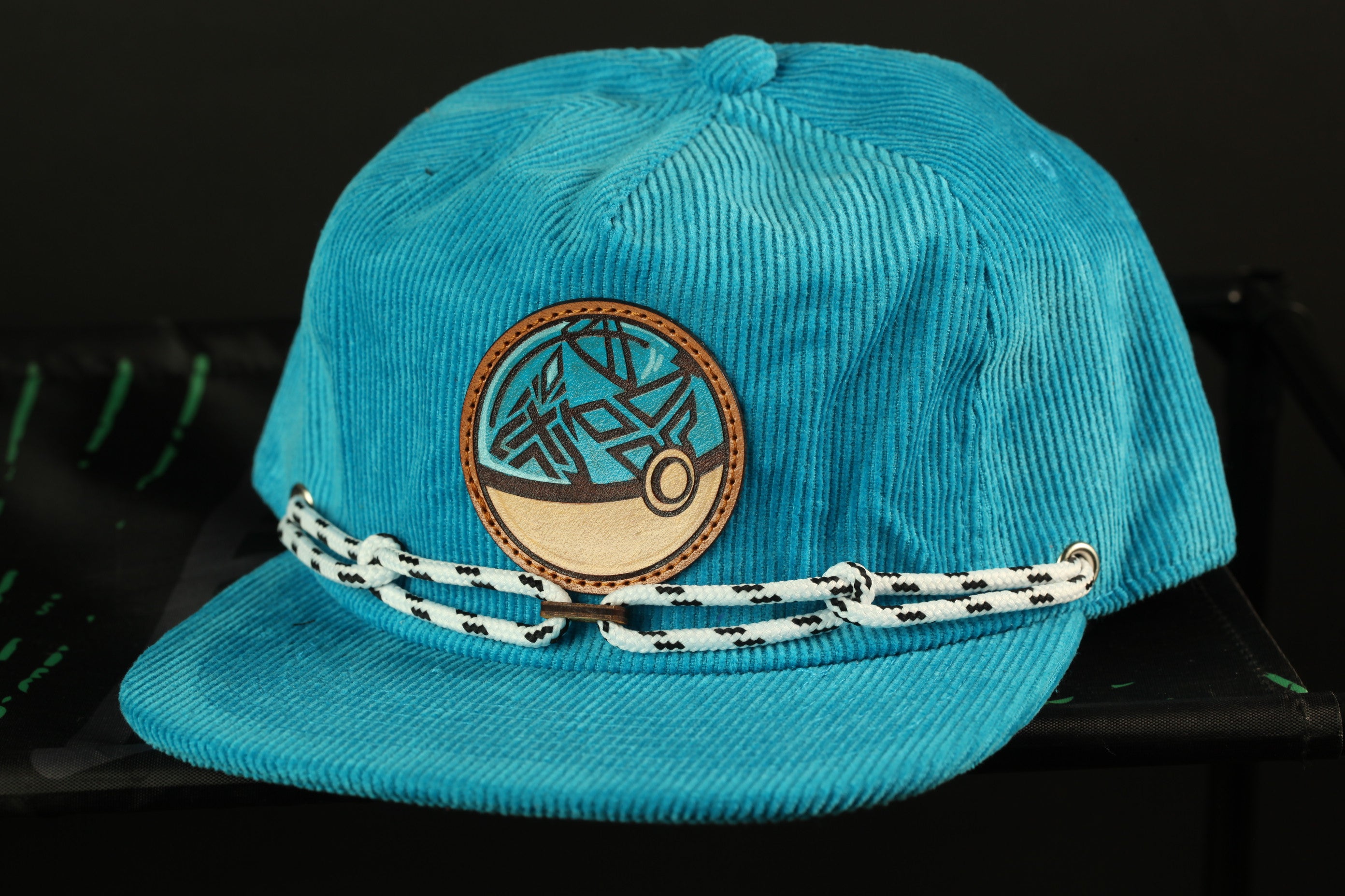 A turquoise baseball cap with a circular patch on the front and a white rope strap.