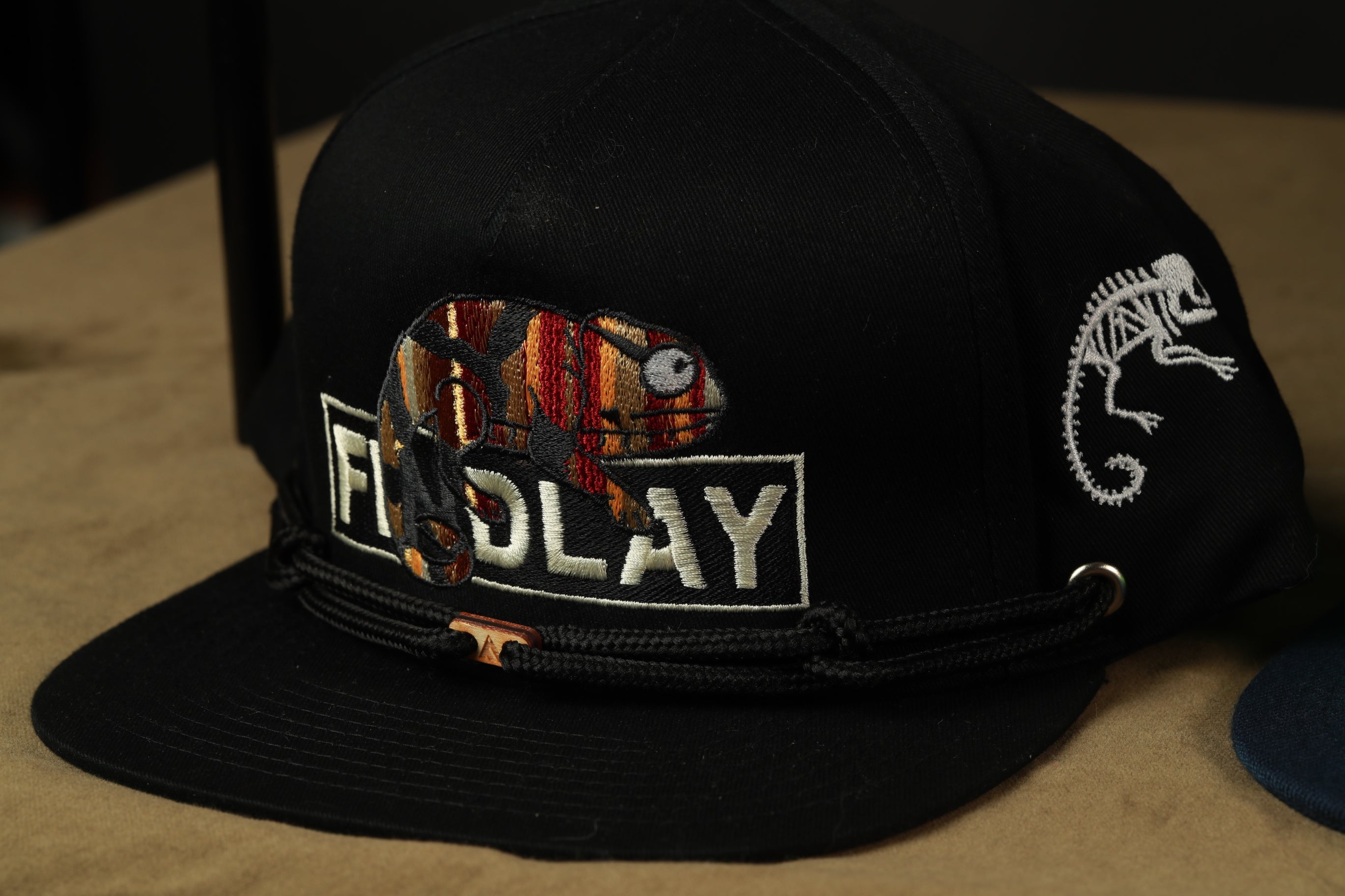 A black baseball cap with a chameleon design and the word "FOLAY" printed on it.