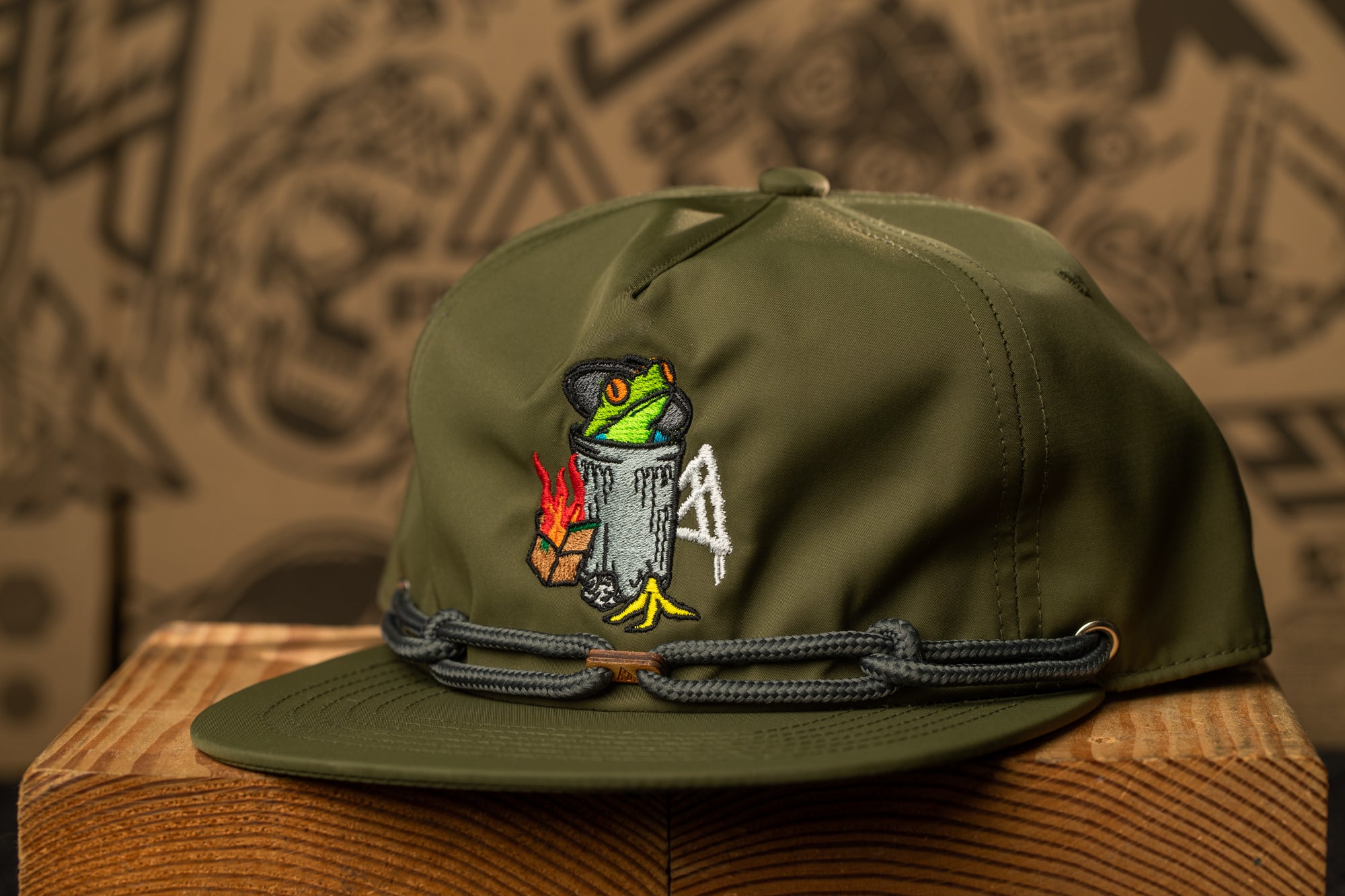A green baseball cap with a frog design and a chain strap, placed on a wooden surface against a background with various abstract patterns.