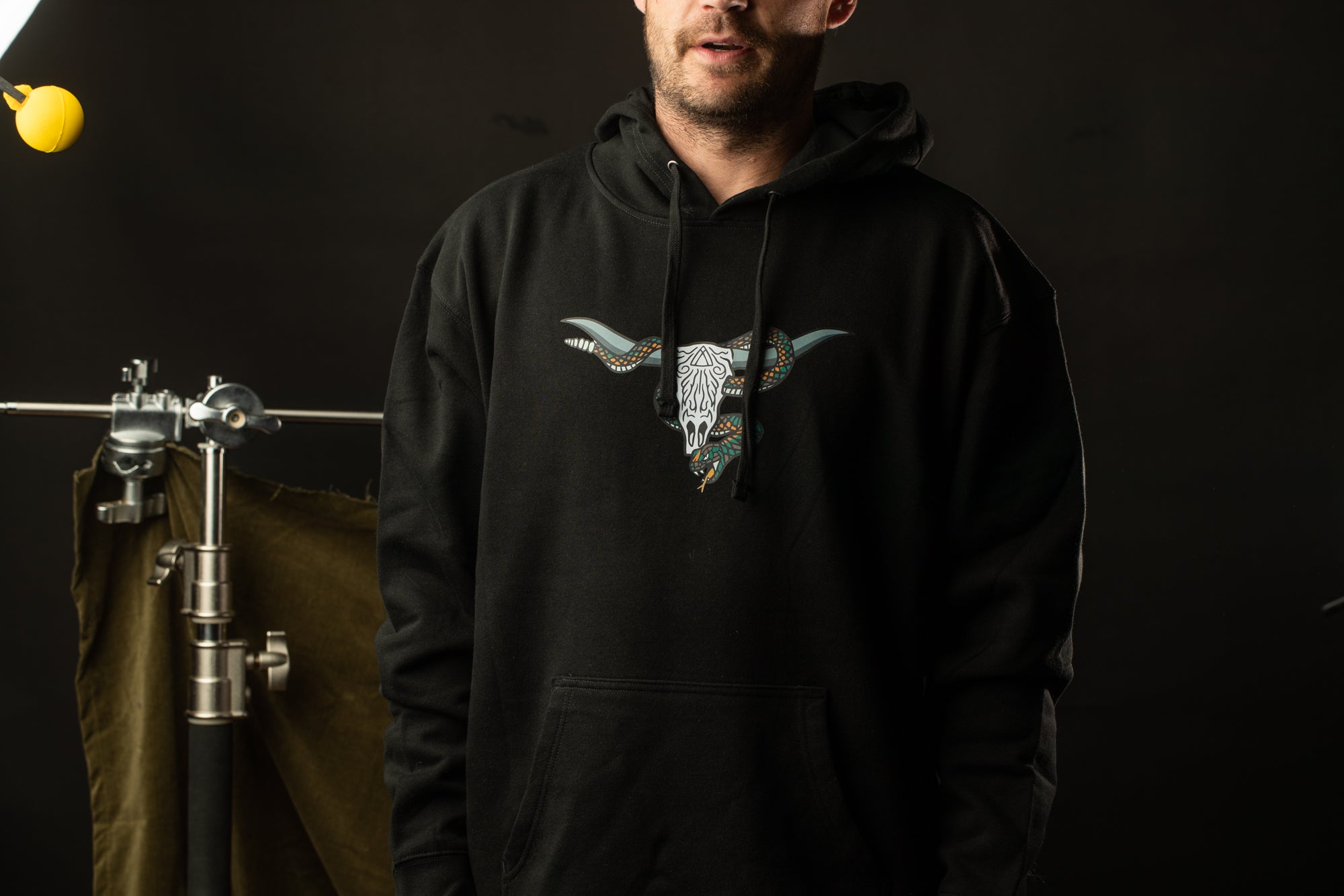A man wearing a black hoodie with a graphic of a bull's head stands in front of a dark background.