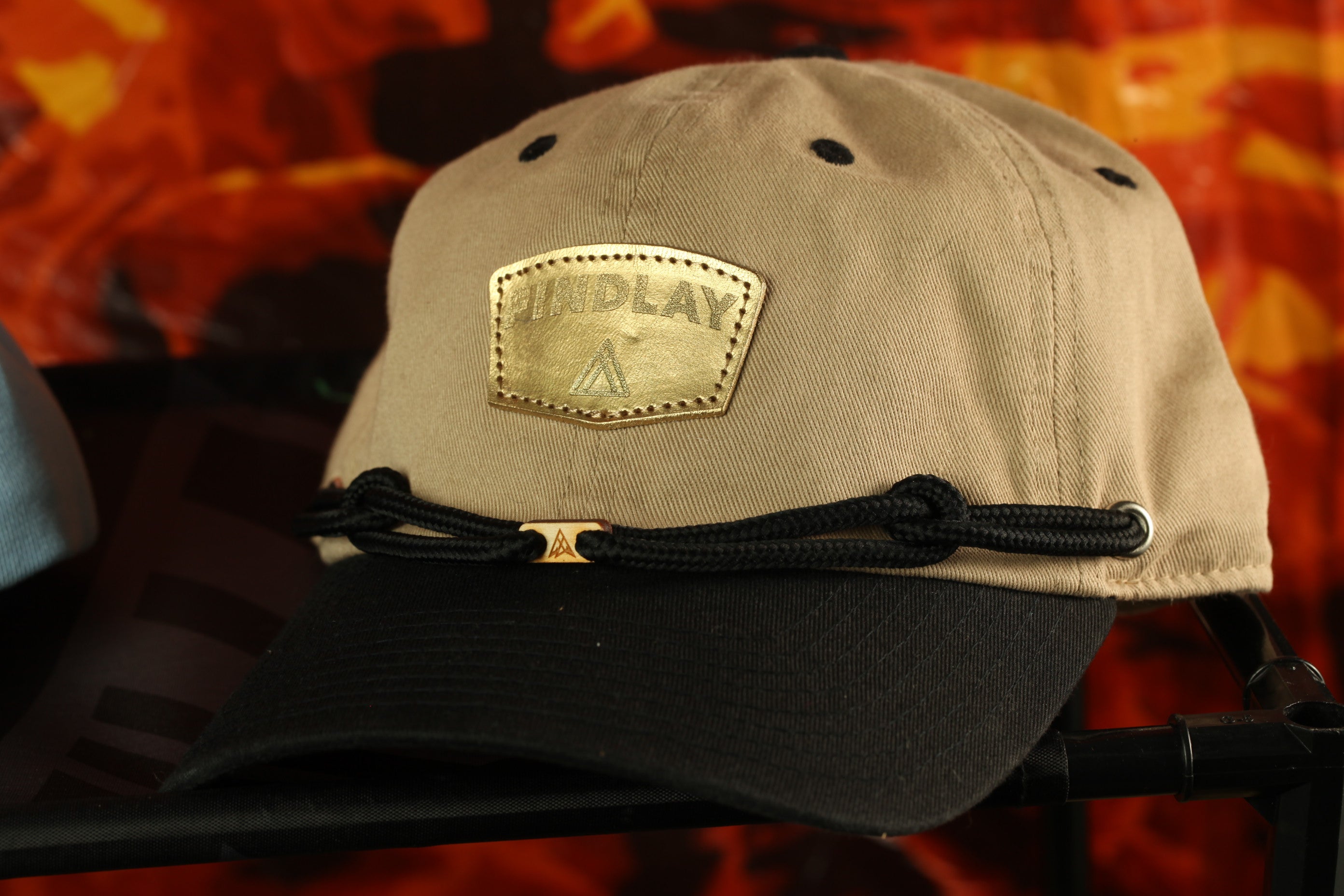 A tan baseball cap with a black brim and a gold-colored patch on the front, displayed on a black stand against a red and orange background.