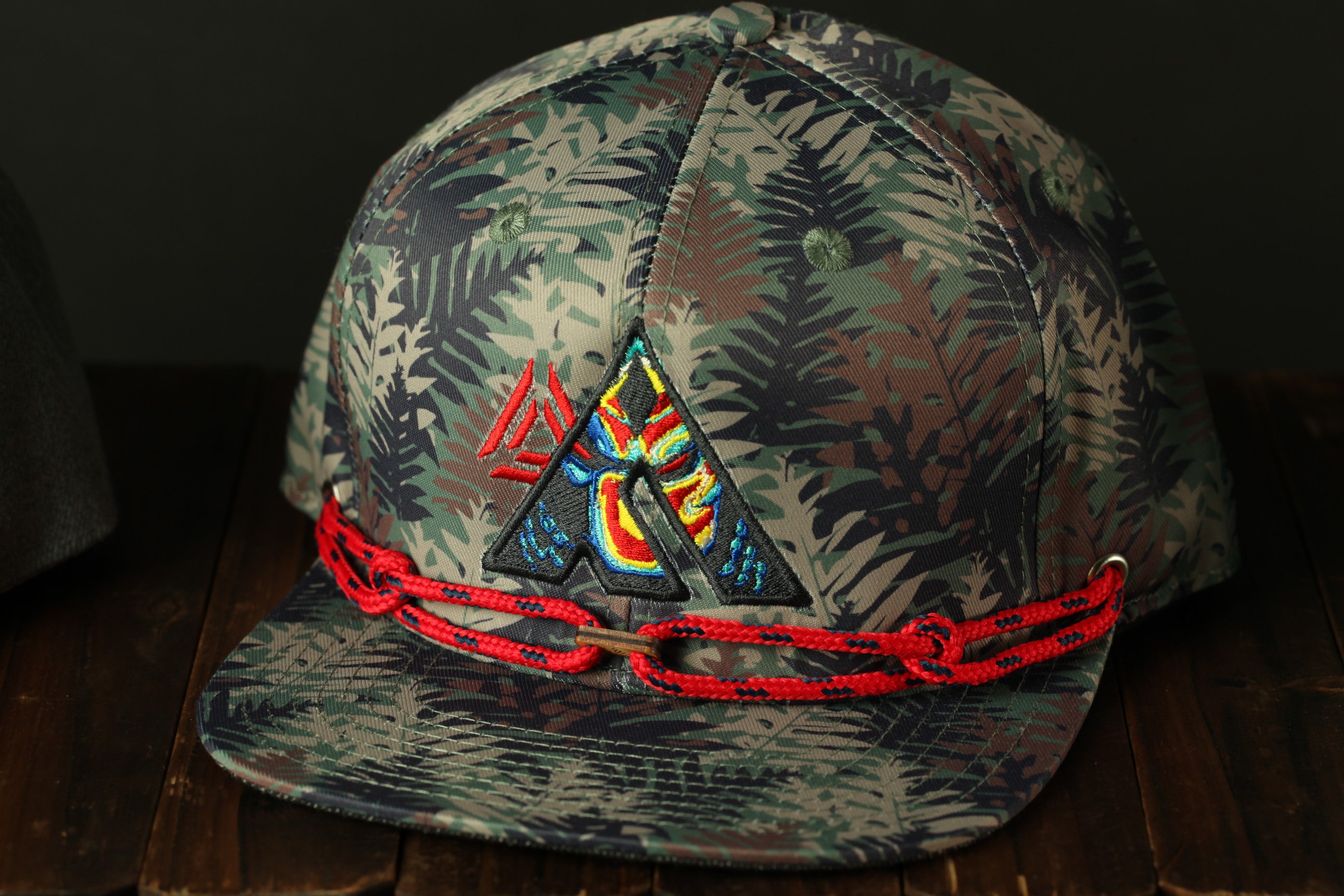 A camouflage baseball cap with a colorful, abstract design on the front, featuring a red chain strap.