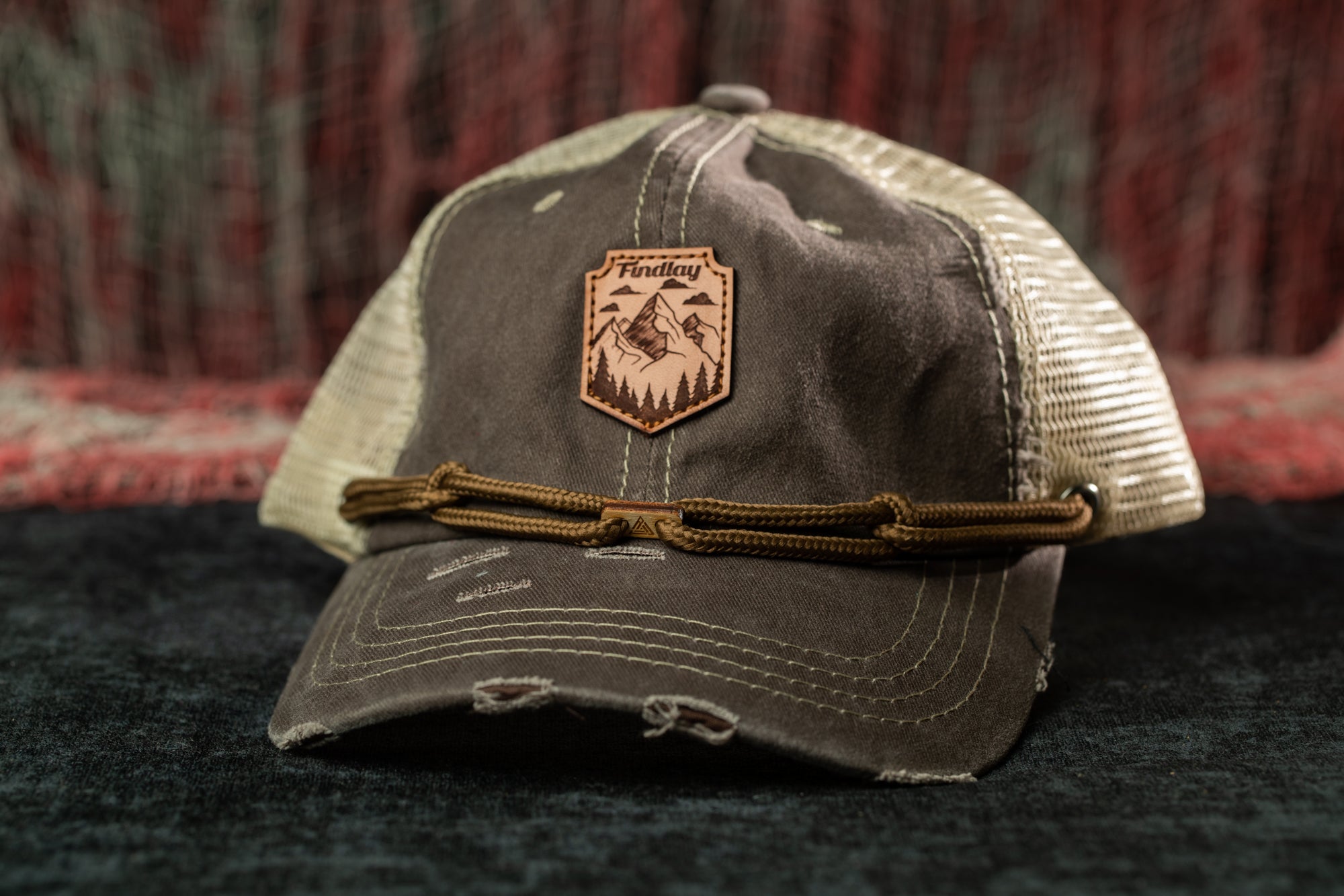 A worn gray baseball cap with a brown leather patch on the front, featuring a mountain landscape design.
