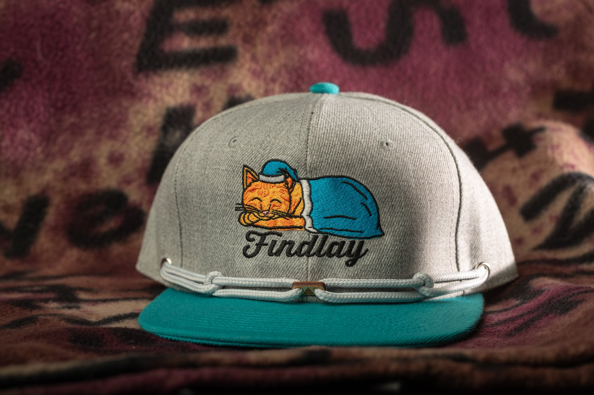 A gray baseball cap with a teal brim and a cat embroidered on the front, sitting on a pink and purple patterned fabric.